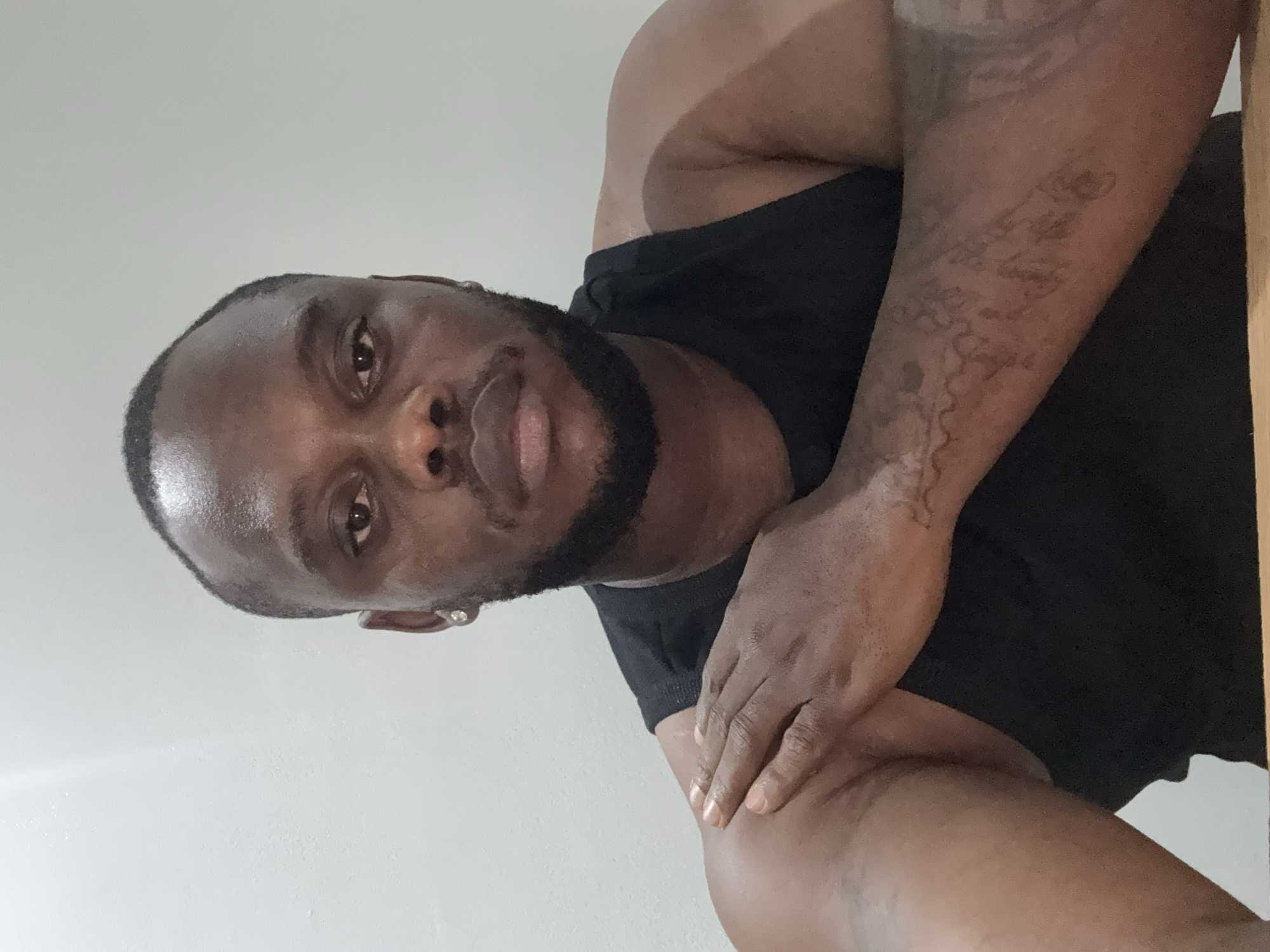 A dark-skinned man is captured in a medium close-up shot, looking directly at the camera with a neutral expression. He is wearing a black sleeveless tank top. His head is slightly tilted, revealing a short, dark haircut and a full, well-groomed beard and mustache. A subtle sheen is visible on his forehead, possibly indicating perspiration. He wears a small, clear stud earring in his left ear. His left arm is bent, crossed over his chest, with his right hand gently resting on his left forearm. Intricate, faded dark tattoos are visible on his left arm and shoulder, appearing as abstract patterns rather than discernible text or clear imagery. The background is a plain, light-colored wall, suggesting an indoor setting. There are no other objects or people visible, and no visible text. The context places this image in Iddo Tudunwada, Nigeria.