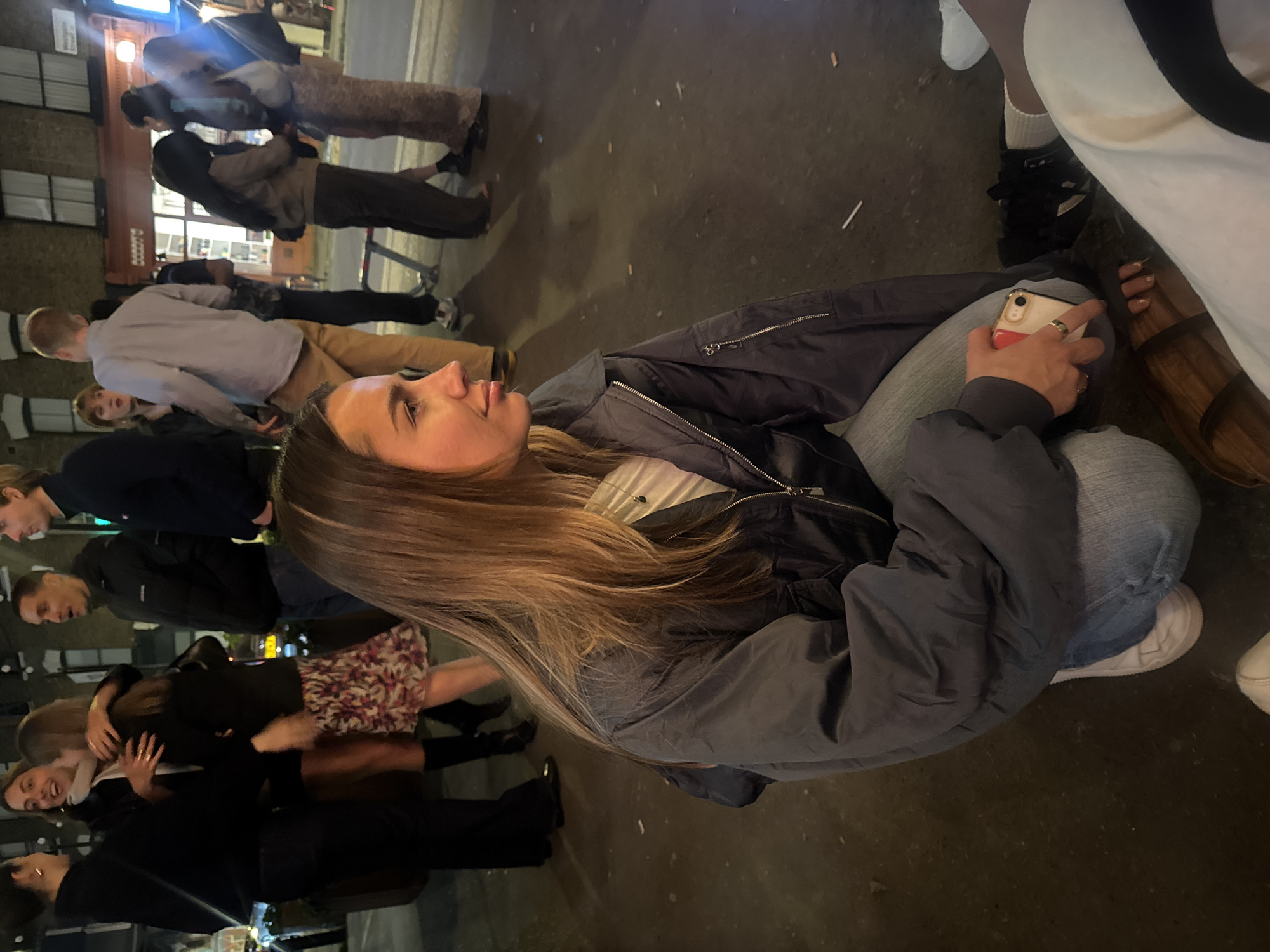 The image captures a candid street scene, likely at night in London, United Kingdom. A young woman is positioned in the foreground, crouching and holding a smartphone in her hand. She is wearing a dark jacket, light jeans, and sneakers. Her hair is long and blonde. In the background, a group of people are standing and interacting. They are dressed in a variety of casual clothing. The environment includes a brick building, a street surface, and faint street lighting.