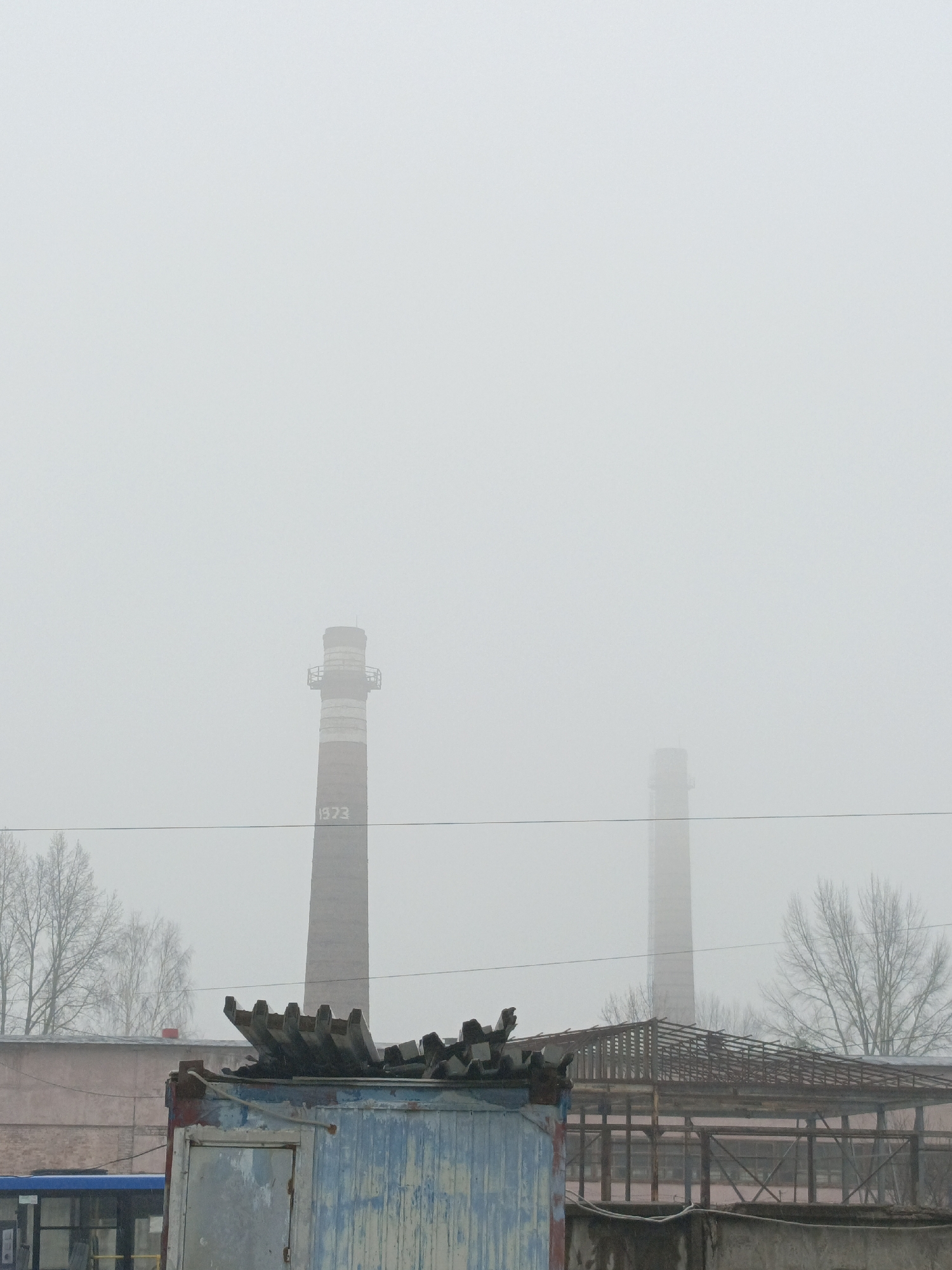 The image shows a foggy, overcast day in Noviy, Russia. In the background, two tall brick smokestacks are visible, with "1973" painted on the closer one. In the foreground, there's a dilapidated structure with a partial blue wall and a collapsed roof. Behind this structure is a pink building and a rusty metal framework of another building. Bare trees stand on either side of the smokestacks. The lower left corner shows a portion of a blue bus. Power lines run horizontally across the image.