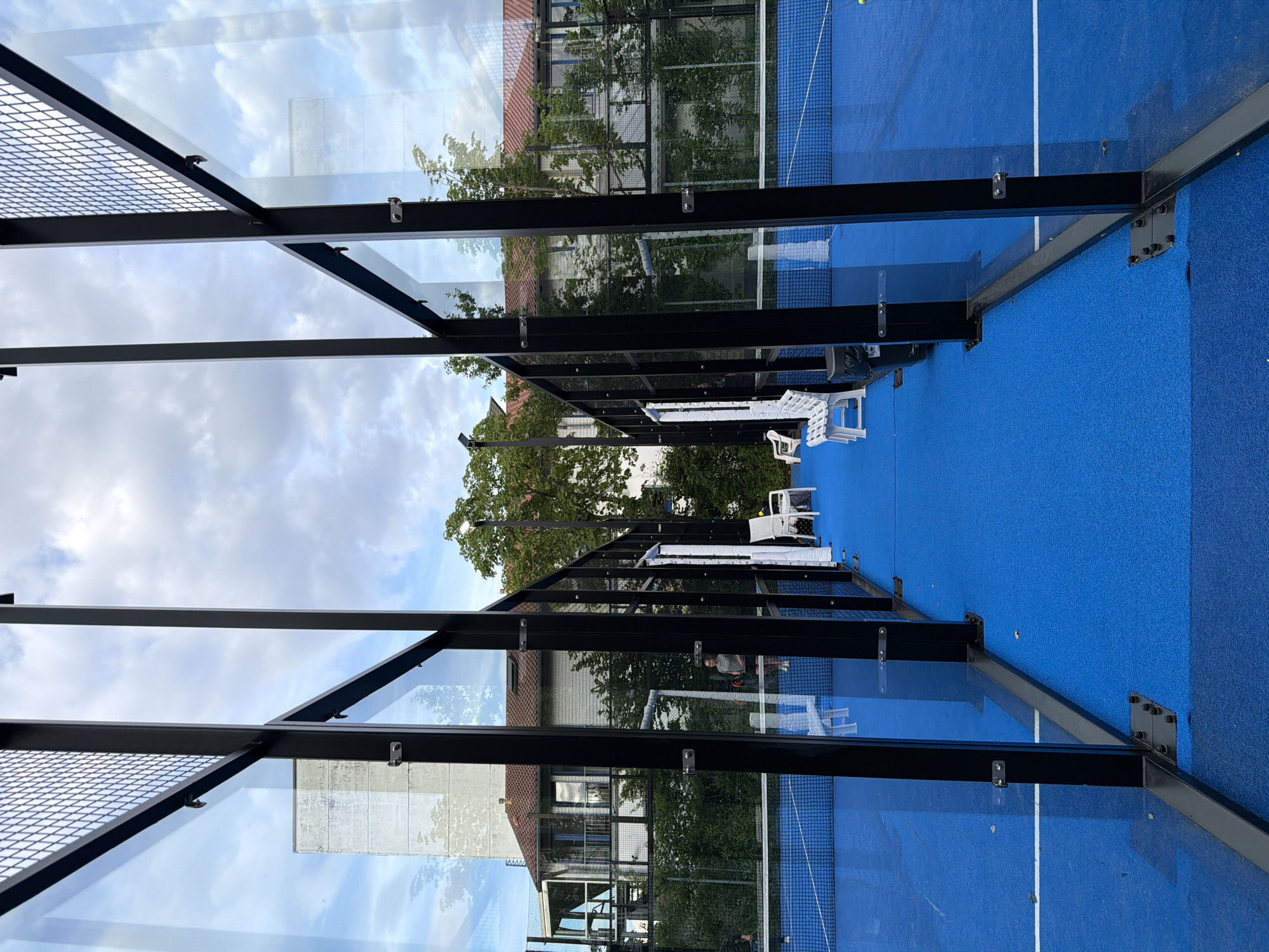 The image captures an outdoor padel tennis court in Vaterstetten, Germany, under a cloudy sky. The court features a bright blue surface with white boundary lines. The court is enclosed by glass walls with black metal frames, and mesh fencing is visible at the top of the enclosures. In the background, buildings with red tiled roofs and trees can be seen. Two individuals are present on the court, engaged in what appears to be a game of padel. One person, wearing a grey t-shirt and shorts, stands near the net, holding a padel racket. Another person, dressed in dark clothing, is partially visible to the left. White plastic benches are positioned along the side of the court. A streetlamp illuminates the area.