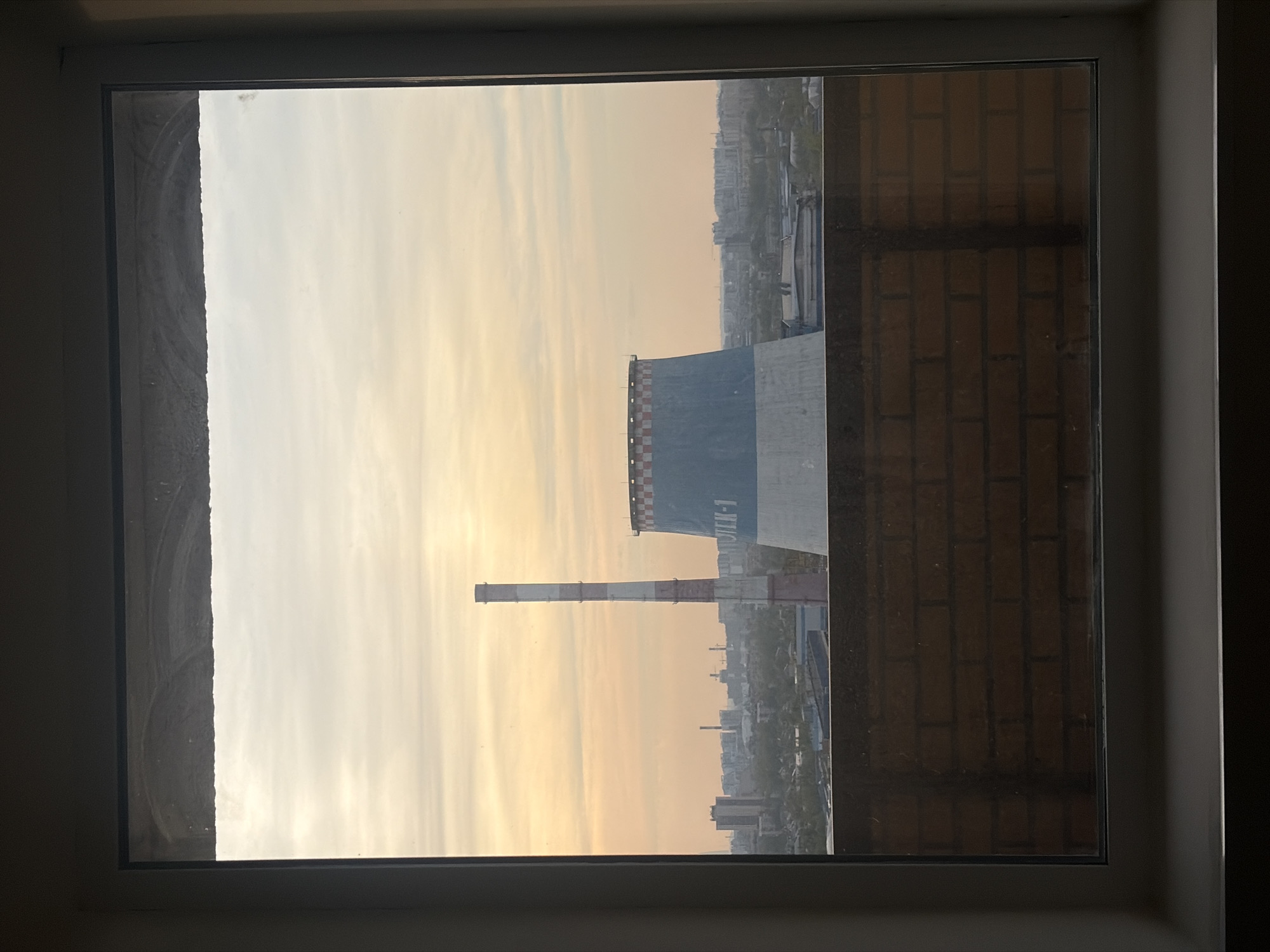 A view through a window depicts a cityscape at dusk or dawn, presumably from a high-rise building. The sky is a blend of pale orange and grey, indicative of either sunrise or sunset. In the foreground is what appears to be a cooling tower of a power plant, painted grey with checkered patterns near the top. A tall chimney, painted with alternating red and white bands, stands nearby. In the distance, various buildings and structures of a city stretch out under the sky. The window frame is visible, adding a sense of depth and perspective to the scene. Given the architecture and the inscription "TЭЦ-1" on the side of the cooling tower, the location is most likely Saint Petersburg, Russia, specifically viewing the "TETS-1" combined heat and power plant.