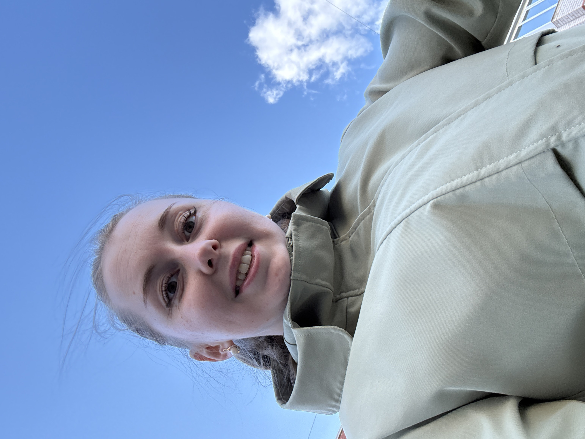 A woman is looking down at the camera from below. She has light skin and blonde hair pulled back. She is wearing a light-colored jacket with a collar. The background is a bright blue sky with a small white cloud. A building is visible in the upper right corner. The city of Unknown, Unknown cannot be determined from this image.