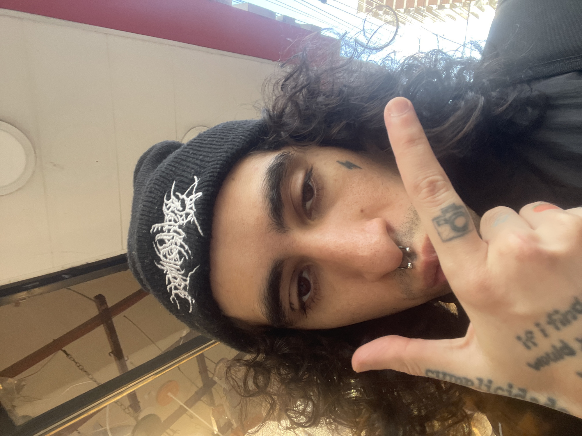 A medium-shot, angled selfie of a young adult male with curly brown hair. He is wearing a black beanie with white gothic-style lettering. He has a small lightning bolt tattoo near his left eyebrow and a septum piercing. The man is extending his right hand towards the camera, index finger pointed up and the thumb extended out, with visible tattoos on his hand. A camera tattoo is visible on the finger with the tip slightly colored with what appears to be a pink marker. Additional tattoos include text which is not completely legible. The location is suggested to be in Buenos Aires, Argentina, with building features visible in the angled shot and reflected in what appears to be glass.