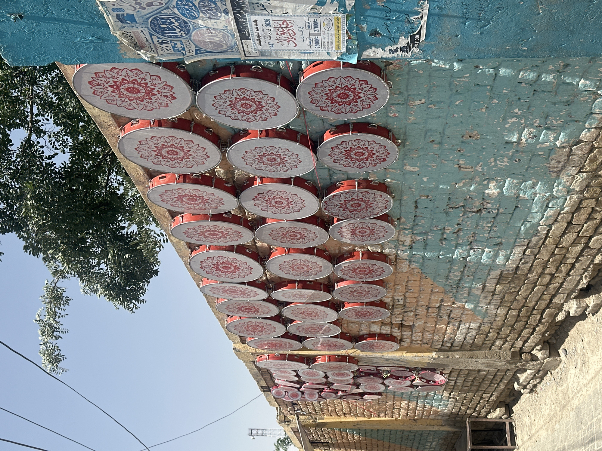 The image captures an outdoor scene, likely a street, adorned with a decorative canopy made of numerous drum-like objects. These round instruments feature red rims and white faces, each intricately decorated with a red mandala pattern. They are arranged in multiple rows, hanging horizontally, with smaller instruments visible further down the display.
Above the drums, a part of a building structure is visible, featuring a blue/teal painted section with peeling paint that reveals underlying brickwork. The right side of the image shows more exposed brick, some of which also has remnants of paint. Posters and flyers are affixed to the upper section of the structure.
To the left, lush green tree foliage is visible against a clear, bright blue sky, indicating a sunny daytime scene. Utility lines stretch across the sky in the lower left. The bright sunlight casts shadows on the drums and the wall.
Visible text on the posters, in Urdu/Arabic, includes the name 'جامعه نجم العلوم للبنات' (Najm al-Uloom Girls' Institute), dates such as '2026' and '1447' (a Hijri year), and financial figures like '25000/=' and '27000/='. Other phrases like 'كلى مجان بشين' (likely Kalli Majan Pishin, relating to the provided location of Pishin, Pakistan) and mentions of 'billing ongoing' suggest announcements or advertisements for the institute. The overall impression is one of festive decoration in an urban or semi-urban setting, possibly for an event or celebration associated with the institute in Pishin, Pakistan.