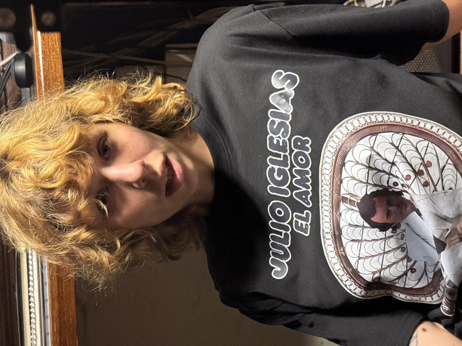 A person with curly, light brown hair is wearing a black t-shirt with a print of Julio Iglesias' name and album title "El Amor" above a circular design featuring the artist. The person is angled towards the camera, with their face visible, including their eyes, nose, and mouth. The background is dark and indistinct, but appears to be an indoor setting. Some wooden furniture is visible behind the person.
