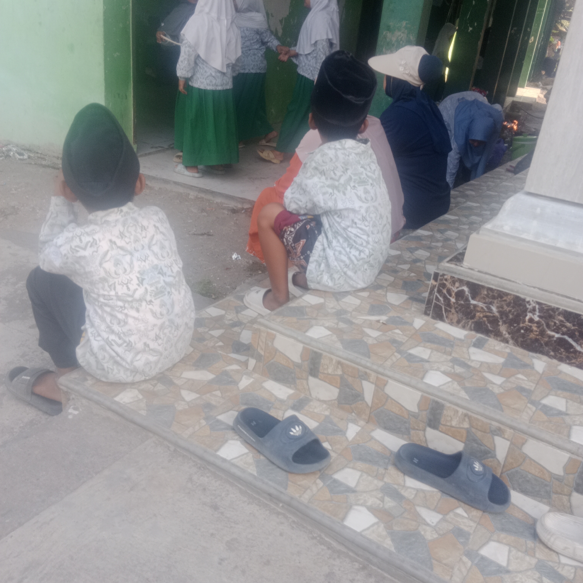 In the city of Sidorejo, Indonesia, two young boys, facing away from the camera, sit on a tiled stairway. The boy on the left, wearing a dark peci and a white patterned shirt, is seated with his legs crossed. The boy on the right, also wearing a peci and a similar patterned shirt, is in a squatting position, his legs tucked underneath him. Between them, on the stairs, is a pair of dark gray slip-on sandals. To the right of the boys, on the same tiled steps, another pair of dark gray slip-on sandals is visible. The background shows a light green wall of a building and further back, other individuals in Islamic attire, including a person wearing a wide-brimmed hat. The ground in front of the stairs is concrete.
