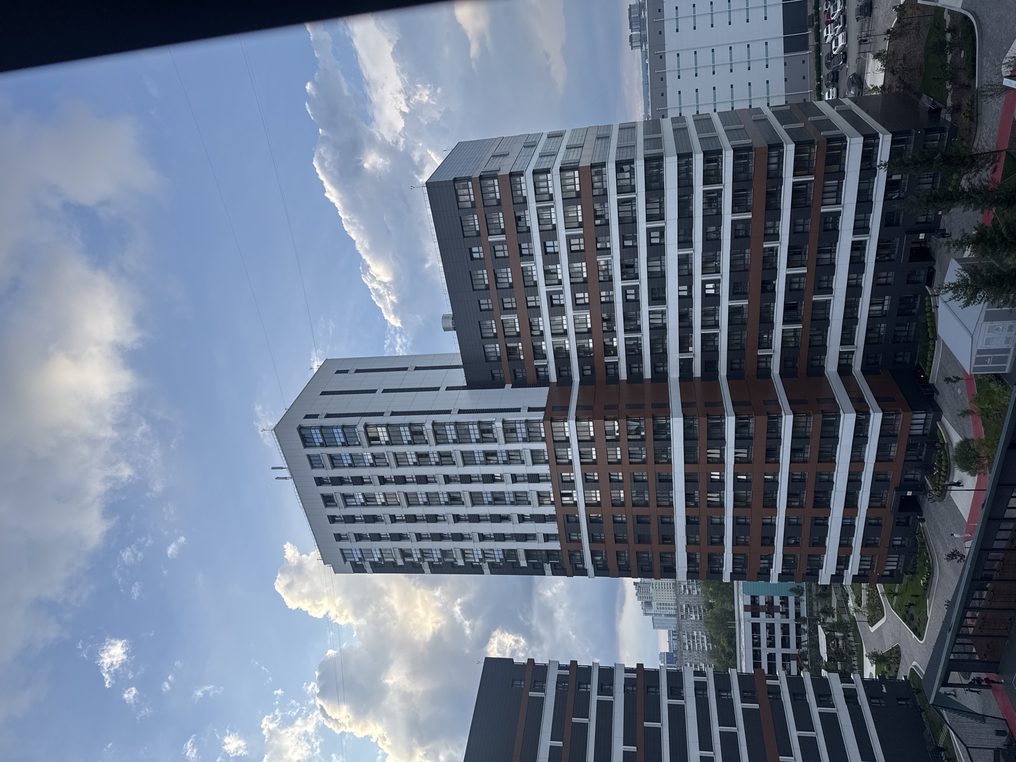 A wide shot captures a modern residential complex in Novosibirsk, Russia, under a cloudy sky. Two tall, multi-story buildings dominate the frame, their facades a mix of dark gray, white, and brown horizontal panels, with numerous rectangular windows. The building on the right is slightly shorter and has a more angular design, while the building on the left is taller and more rectangular. In the foreground, a dark railing partially obstructs the view, suggesting the photo was taken from an elevated position, possibly a balcony. Below the buildings, an open courtyard features paved pathways, green grassy areas, and scattered trees. In the distance, other buildings, including what appears to be a large warehouse or industrial facility with a light-colored facade and a grid pattern, are visible. A parking lot with several cars is situated near the industrial building. The sky is a mix of blue and white clouds, with some sunlight breaking through. A few individuals are discernible in the courtyard, walking along the paths. The overall impression is of a new urban development with a contemporary architectural style.