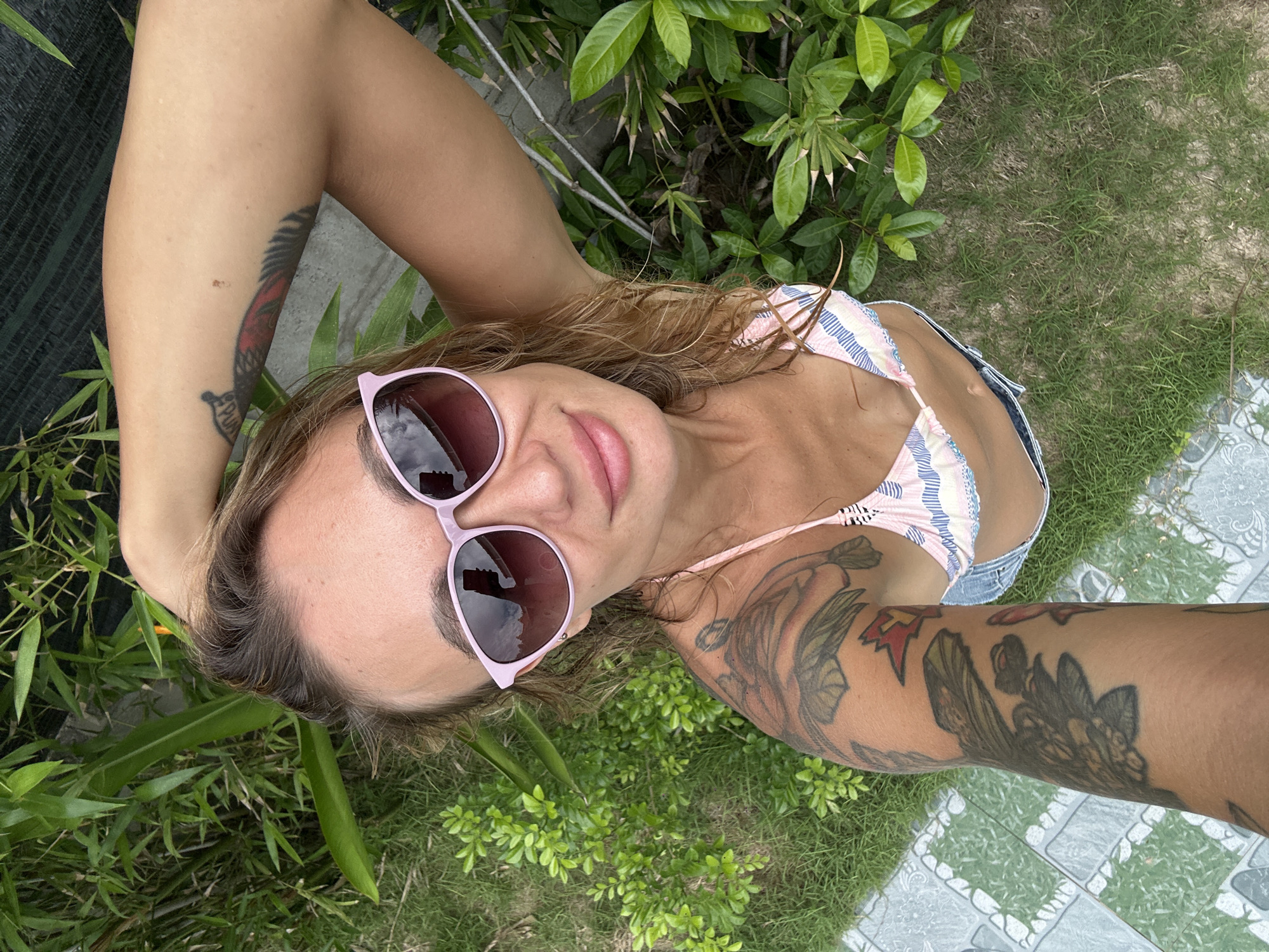 A close-up, high-angle portrait captures a person relaxing outdoors in Mũi Né, Vietnam. The subject, likely female, is seen from above, lying down or reclining, with her head centered and looking upwards at the camera. She wears light pink, reflective sunglasses and a pastel-striped bikini top. Her lips are slightly parted in a relaxed, subtle smile.
Both arms are extensively adorned with tattoos. Her left arm (on the viewer's right) features a prominent design with red and dark elements, including a stylized feather and the clearly legible word "PUN" near the forearm, along with a red star symbol on the inner upper arm. Her right arm (on the viewer's left) displays a large, intricate sleeve of dark, organic, possibly floral or foliage patterns. Her medium-brown hair is loosely spread around her head.
The background consists of lush green vegetation, including various leafy plants and grass, with a grey and white patterned tiled surface visible along the edges. This suggests an outdoor garden, patio, or resort setting. The scene is brightly lit by natural daylight, indicating sunny and good weather conditions.
