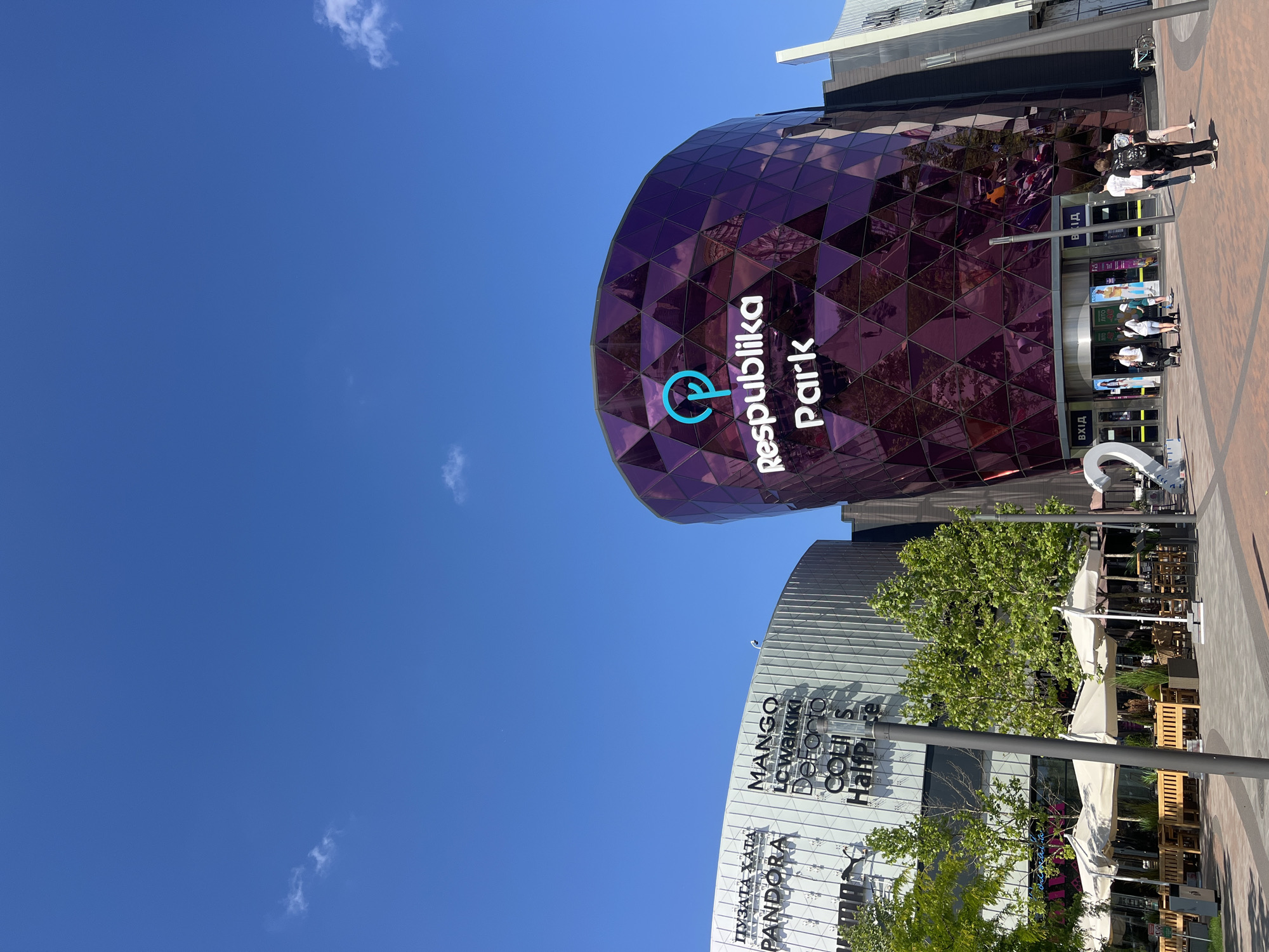 The image shows a clear, bright blue sky with a few wispy clouds. Below the sky, two modern buildings are visible. The building on the right is a striking purple structure with a faceted, geometric facade. It features a large, white logo that reads "Respublika Park" with a blue circular symbol above it. Below the logo, there are entrances with the word "ВХІД" (Entrance) in Cyrillic. Several people are walking near the entrance.
To the left of the purple building is a lighter-colored building with a textured facade. This building displays various brand names in large, dark lettering, including "ПУЗАТА ХАТА," "PANDORA," "MANGO," "LC Waikiki," "DeFacto," "Colins," and "HalfPrice." In front of these buildings, there is an outdoor seating area with tables, benches, and umbrellas, suggesting a cafe or restaurant. Several trees with green foliage are planted in front of the buildings. The ground is paved with reddish-brown bricks. The overall scene appears to be an outdoor plaza in a commercial area. The signs indicate that this is the Respublika Park shopping center, located in Kyiv, Ukraine.