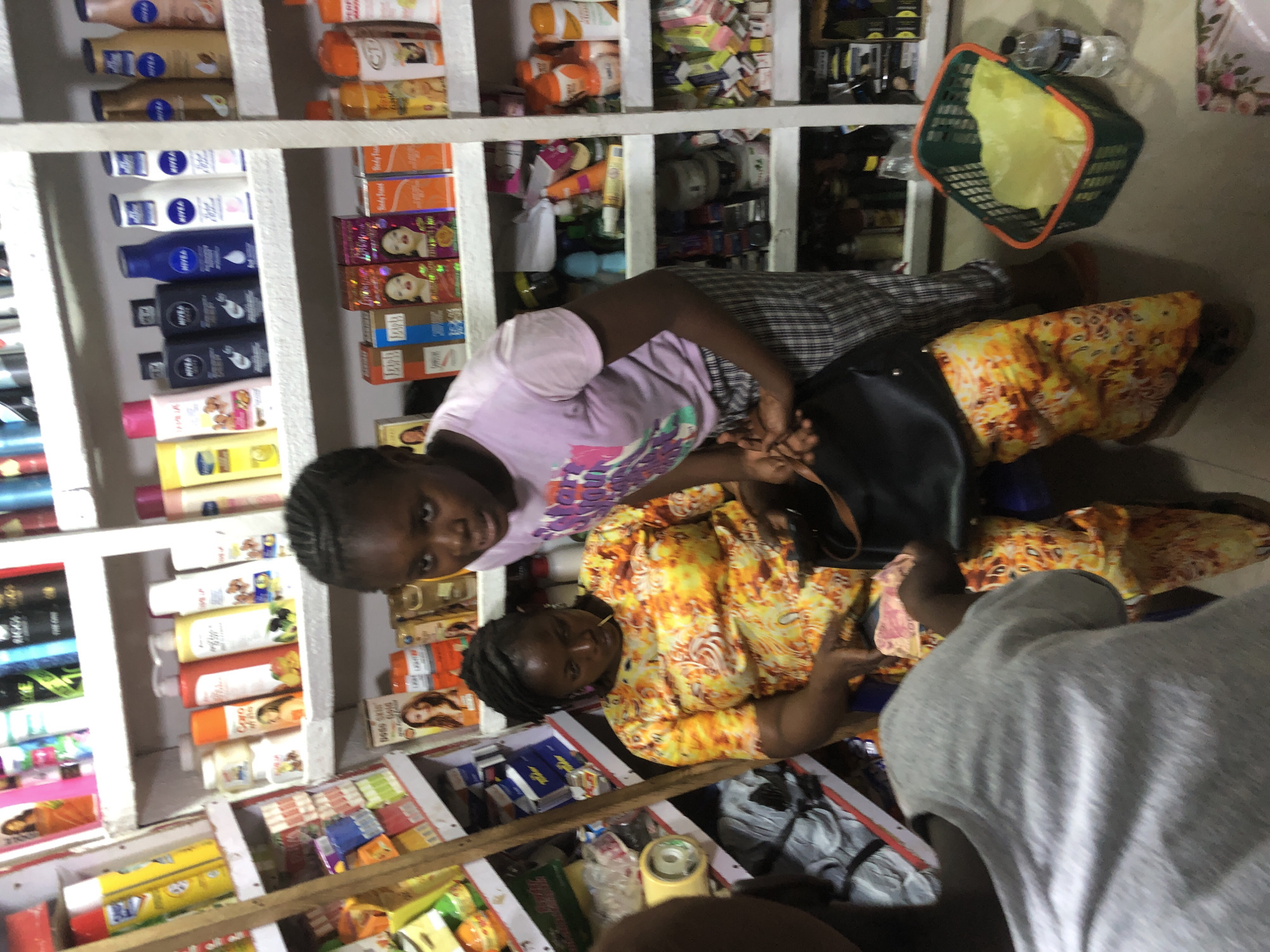 A medium shot captures an interior scene of a store. A young Black female wearing a pink t-shirt and plaid pants stands to the right, looking slightly off-camera to the right. To her left, another Black female sits, wearing a bright yellow and orange patterned dress. She is looking down at her hands, which appear to be holding cash, and a dark purse is slung over her left shoulder. The background is filled with shelves stocked with various retail items, including bottles of lotions, shampoos, and packaged goods, consistent with a small convenience store or pharmacy. The lighting is bright, suggesting artificial interior lighting.