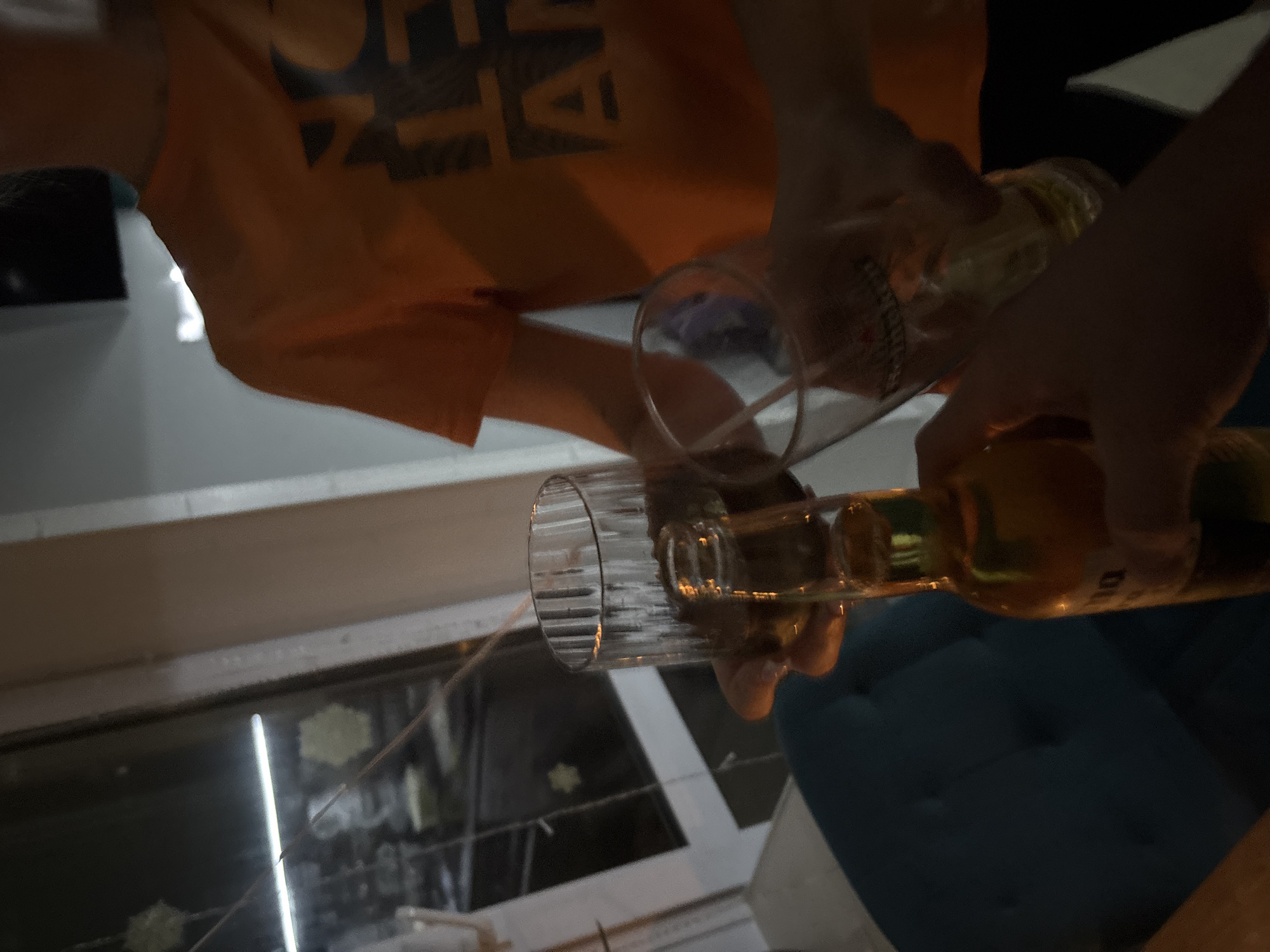 A person wearing an orange shirt is pouring beer from a brown bottle into two glass cups. The location appears to be indoors, based on the window in the background. The image may be in Perm, Russia.