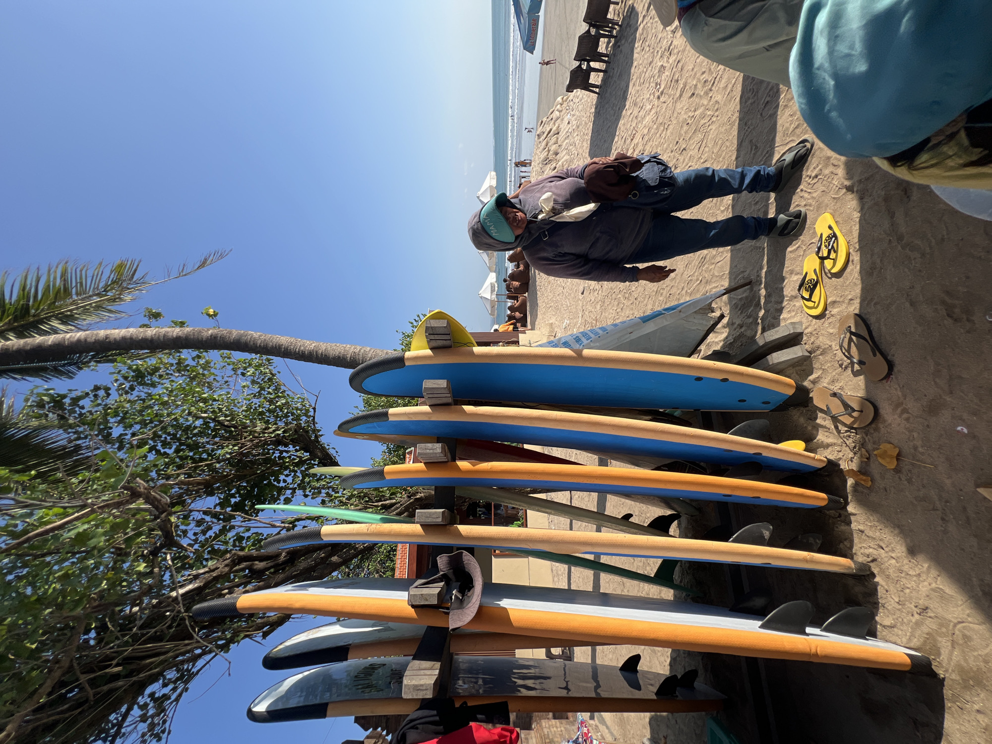 In Kuta, Indonesia, on a sunny beach, a woman stands by a rack of surfboards. She is wearing a gray hooded sweatshirt, blue jeans, and a light blue head covering. She carries a dark blue bag and a brown garment slung over her shoulder. Several surfboards of varying colors, including orange, blue, and gray, are propped upright in a wooden rack on the sand. In the background, the ocean is visible with gentle waves and a few people swimming. Beach chairs and umbrellas are scattered on the sand. The scene is under a clear blue sky with palm trees and other vegetation to the left.