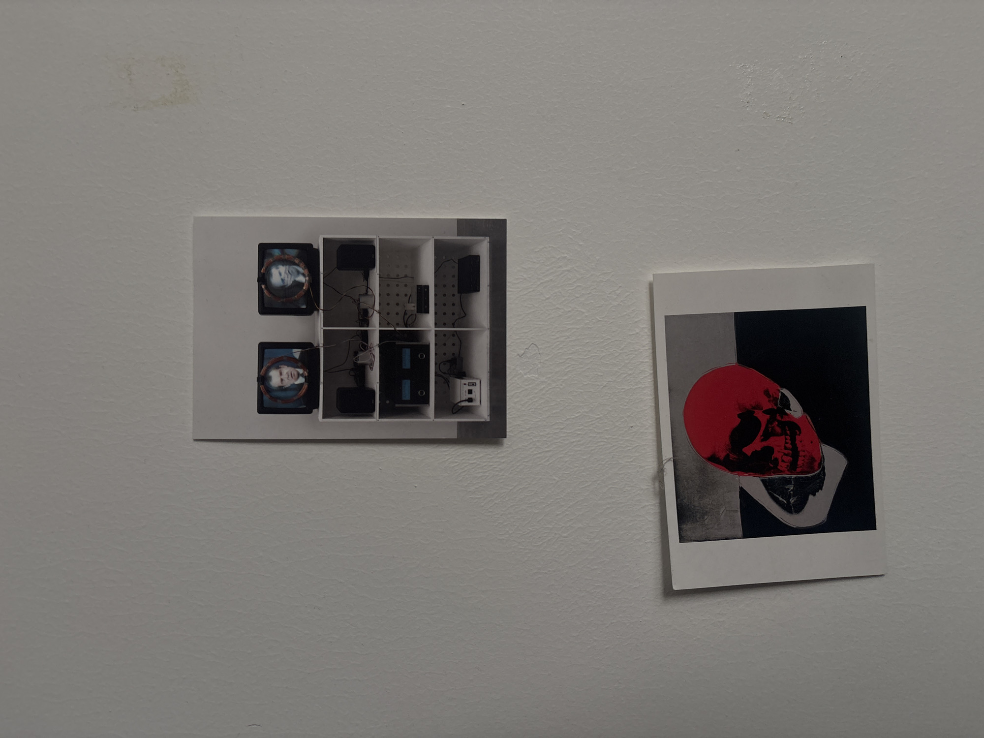 Two photographs are affixed to a white wall, presumably inside a building.
On the left, the photograph depicts a white, compartmentalized structure containing electronic equipment. Two round screens, each displaying a person's face, are visible. Wires connect the components.
To the right, another photograph shows a skull graphic in grayscale with a bright red area covering the skull's top.
The environment is plain with a white wall. The location of San Bernardino Tlaxcalancingo, Mexico, is not discernible from the image.