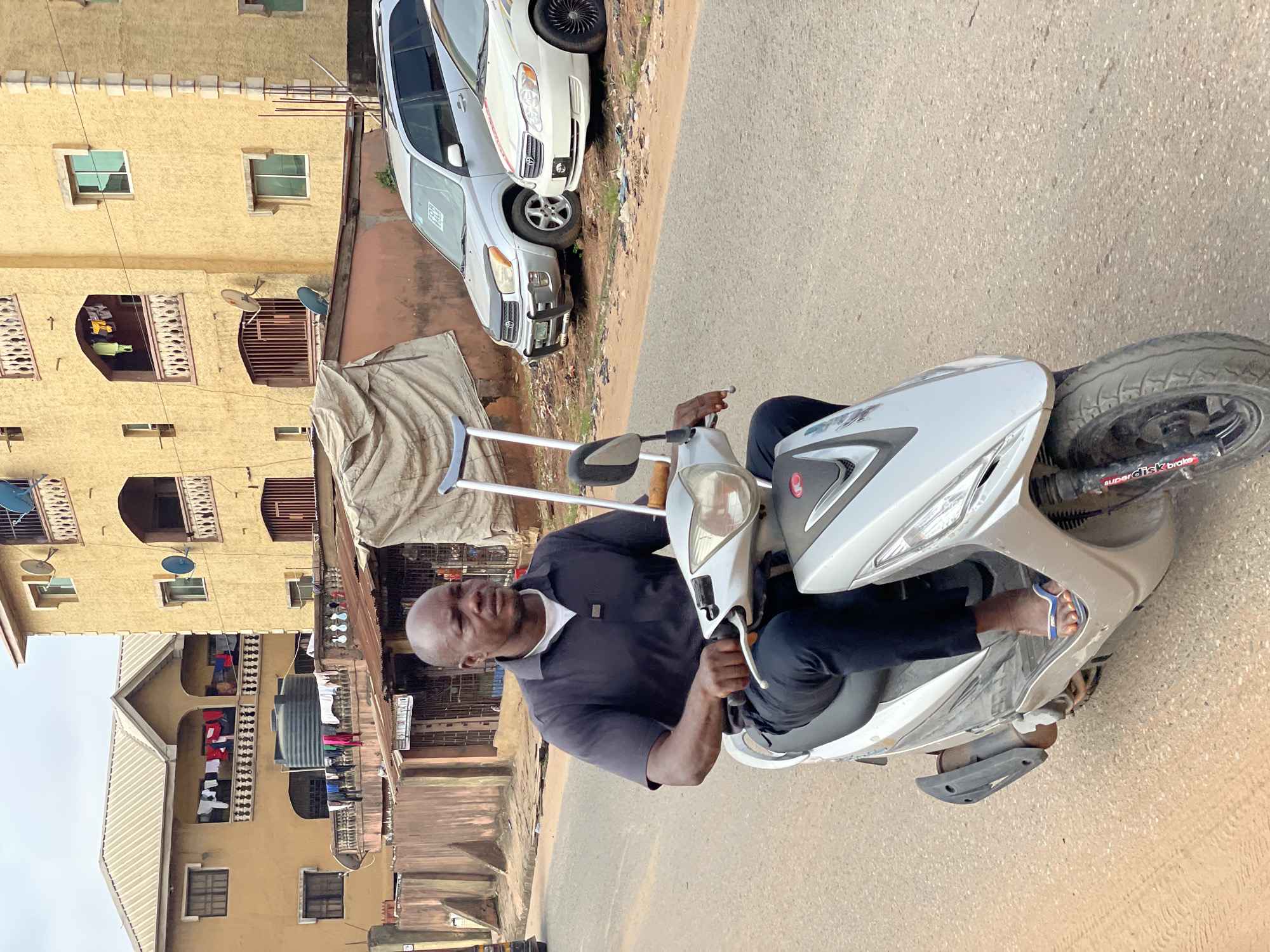 A man rides a silver scooter on a paved road. He wears a navy polo shirt and dark pants, with flip-flops on his feet. The scooter is positioned at an angle, the front tire near the upper-right corner. Buildings line the left side of the street, revealing multi-story apartment blocks with open balconies and laundry hanging out to dry. Vehicles are parked near the building, with a tarp-covered structure between the cars and the buildings. The location is identified as Nkpor, Nigeria.