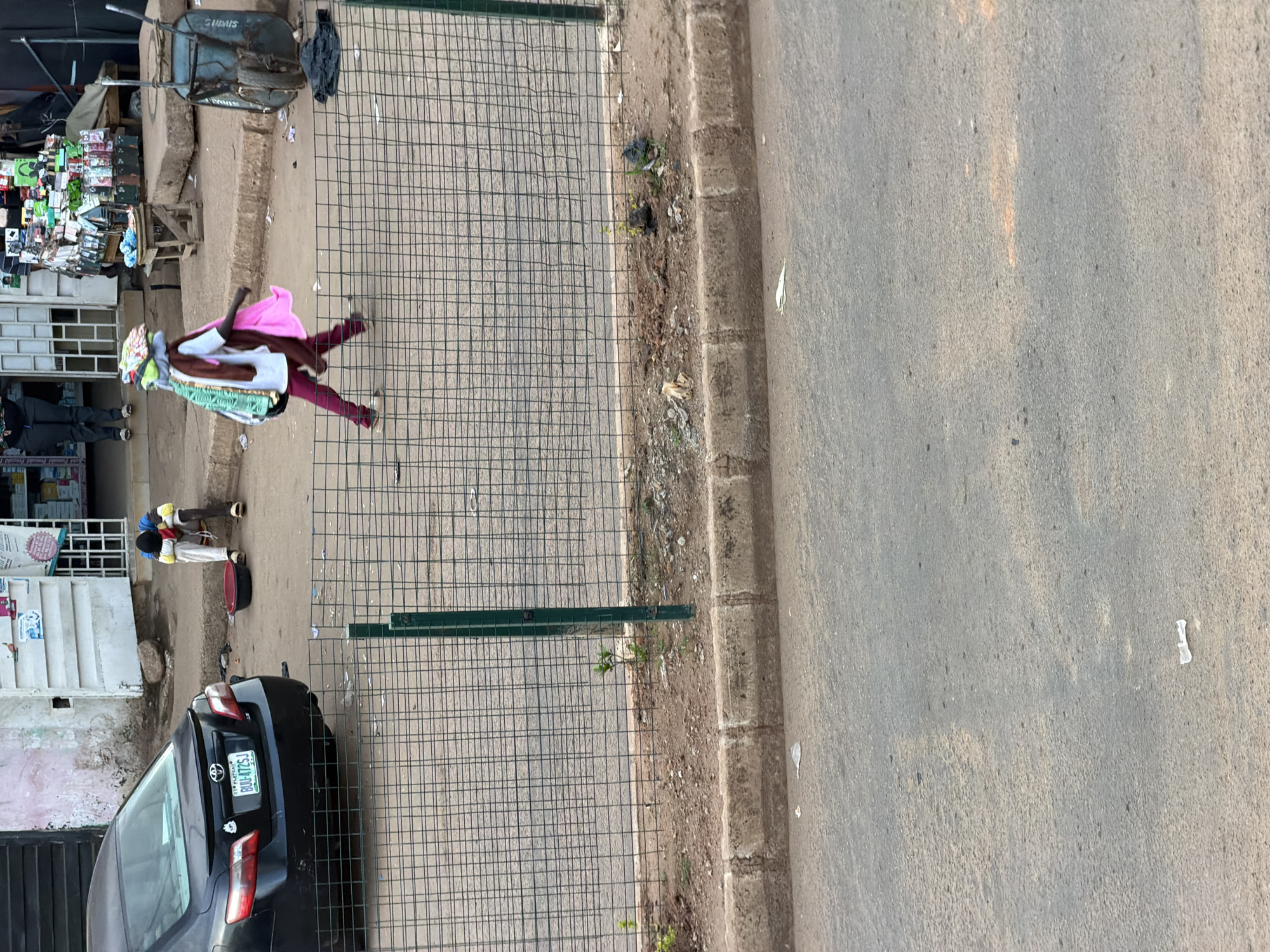 This is a vertical, eye-level shot taken from a sidewalk looking across a street and down a street. On the left side of the frame, a street market is visible. Several vendors are present, with their stalls lined up along the street. One vendor, a woman, is carrying a large stack of fabric items on her head and shoulders. She is wearing a pink headscarf and a white and maroon outfit. Behind her, another person in a dark shirt and pants is walking away. Further down the street, two children are standing near a black car. They appear to be wearing white and yellow clothing. The car has a Nigerian license plate that reads "BOU47ZSJ". The overall scene depicts a bustling street market in a Nigerian town. The time of day appears to be late afternoon or early evening, as indicated by the long shadows cast by the objects and the warm lighting. The weather seems to be fair and dry.