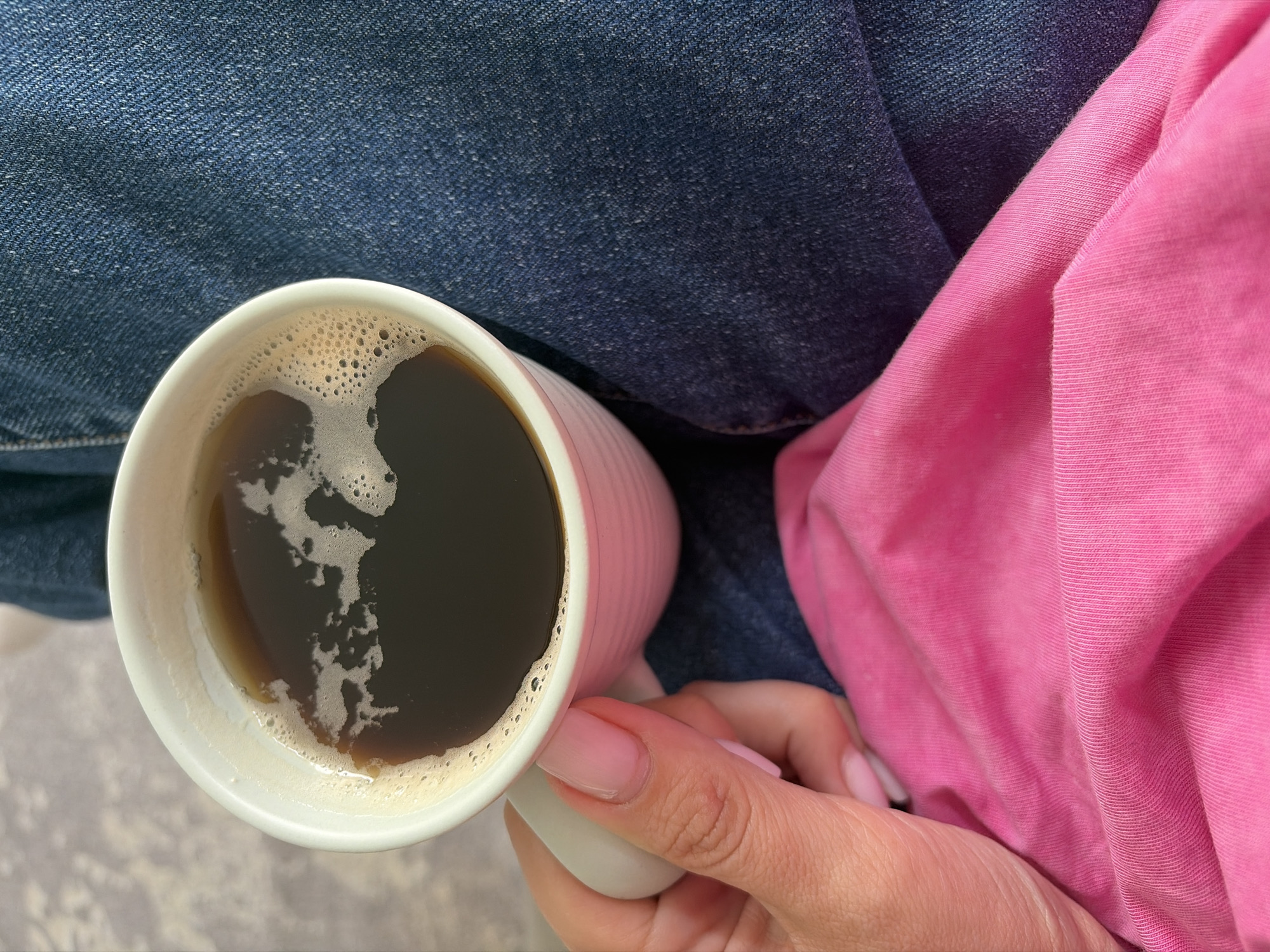 This is a close-up shot of a person holding a white mug of dark coffee. The mug has a ribbed texture. The coffee has a frothy top with some bubbles. The person's hand, with light pink nail polish, is visible holding the mug. The background consists of dark blue denim and a bright pink fabric, which appears to be clothing. The lighting suggests it is daytime, and the scene is casual and comfortable. There is no visible text in the image. The location context is Sochi, Russia.
