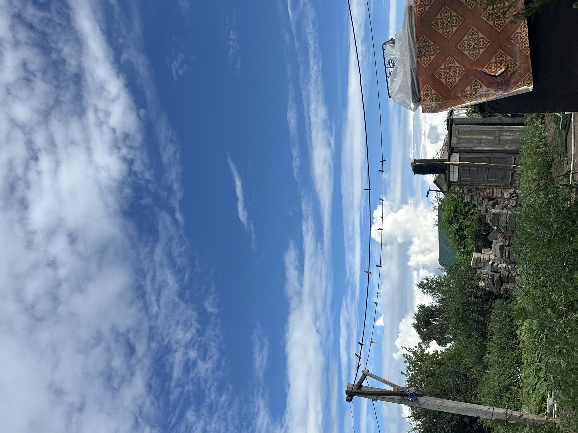 The image depicts a rural scene under a bright blue sky with scattered white clouds. In the foreground, a wooden pole supports a clothesline with several clothespins. To the left of the pole is lush green vegetation, and to the right, a small dilapidated structure with a dark-colored water tank on top and a ladder leaning against it is visible. Behind this structure, a pile of bricks and stones is present. Further in the background, trees and a building with a green roof can be seen. In the far right of the image, a large object covered with a patterned material and clear plastic is partially visible. This scene appears to be in a residential area in Tobyl, Kazakhstan, possibly a backyard or garden. There are no people visible in the image.