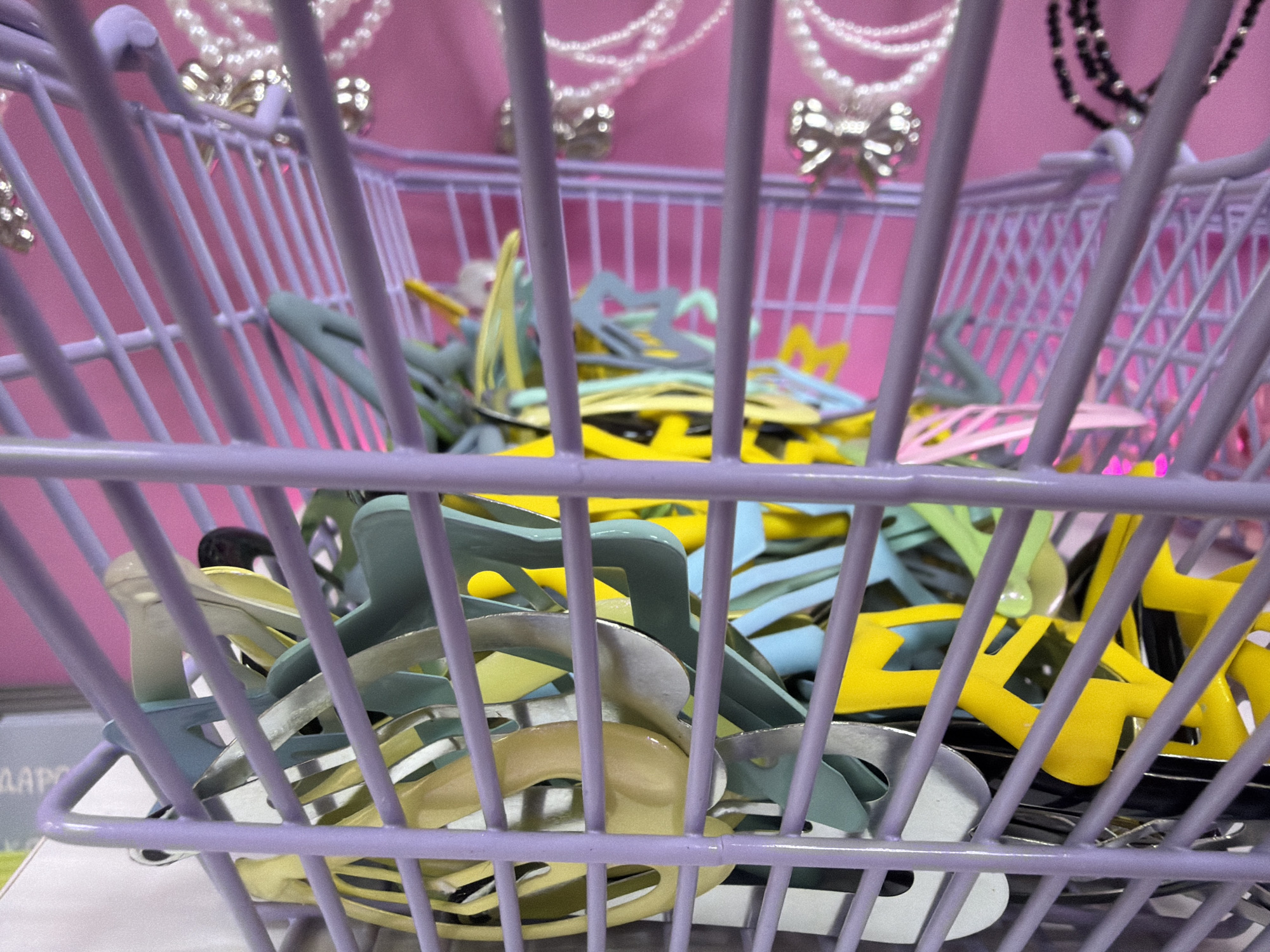 A close-up shot taken indoors shows a small, lavender-colored wire shopping basket filled with an assortment of colorful hair clips. The clips are predominantly in shades of yellow, blue, silver, and green. The basket is situated against a backdrop of pink. The top of the frame shows necklaces with pendants, but without enough resolution to make out much detail.