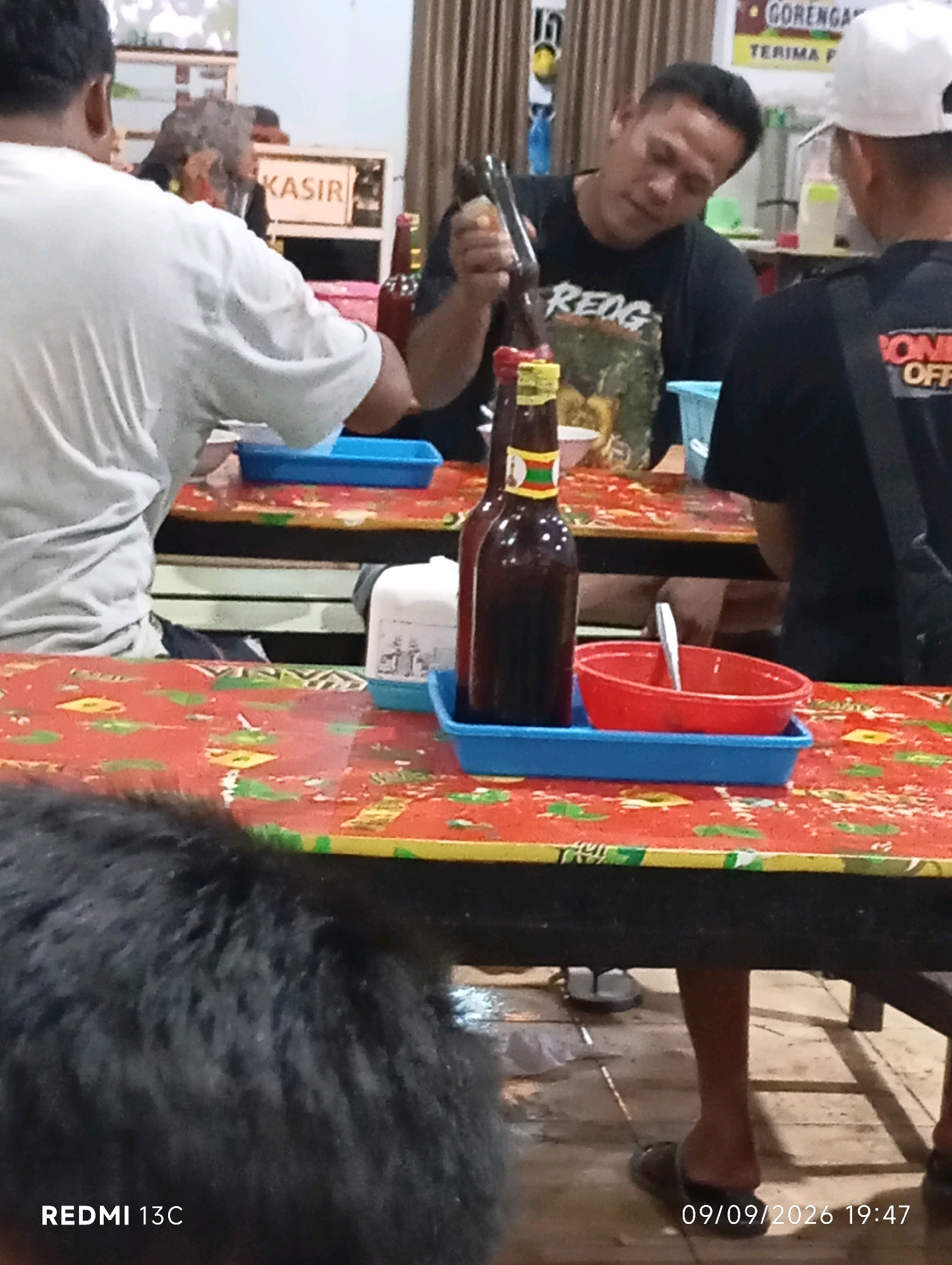 In a dimly lit, casual dining establishment in Wuluhan, Indonesia, two men are seated at a table. The man on the left, facing away from the camera, wears a white t-shirt. The man on the right, also facing away, is dressed in a black t-shirt with a graphic design and a white baseball cap. He is holding a bottle of dark liquid, possibly soy sauce, and appears to be pouring it.
In front of them, on a table covered with a red cloth featuring a colorful pattern, are several items. Two bottles of dark liquid, one with a yellow and green label, are prominent. A blue tray holds a red bowl with a spoon inside. To the right, a white dispenser with a blue lid sits next to another blue tray. In the background, a sign with the word "KASIR" (cashier) is visible. The floor is wooden.
A portion of another person's dark, furry head is visible in the foreground, obscuring the lower part of the table. The bottom right corner of the image displays the text "REDMI 13C" and the date "09/09/2026 19:47". The overall ambiance suggests a relaxed, informal setting.