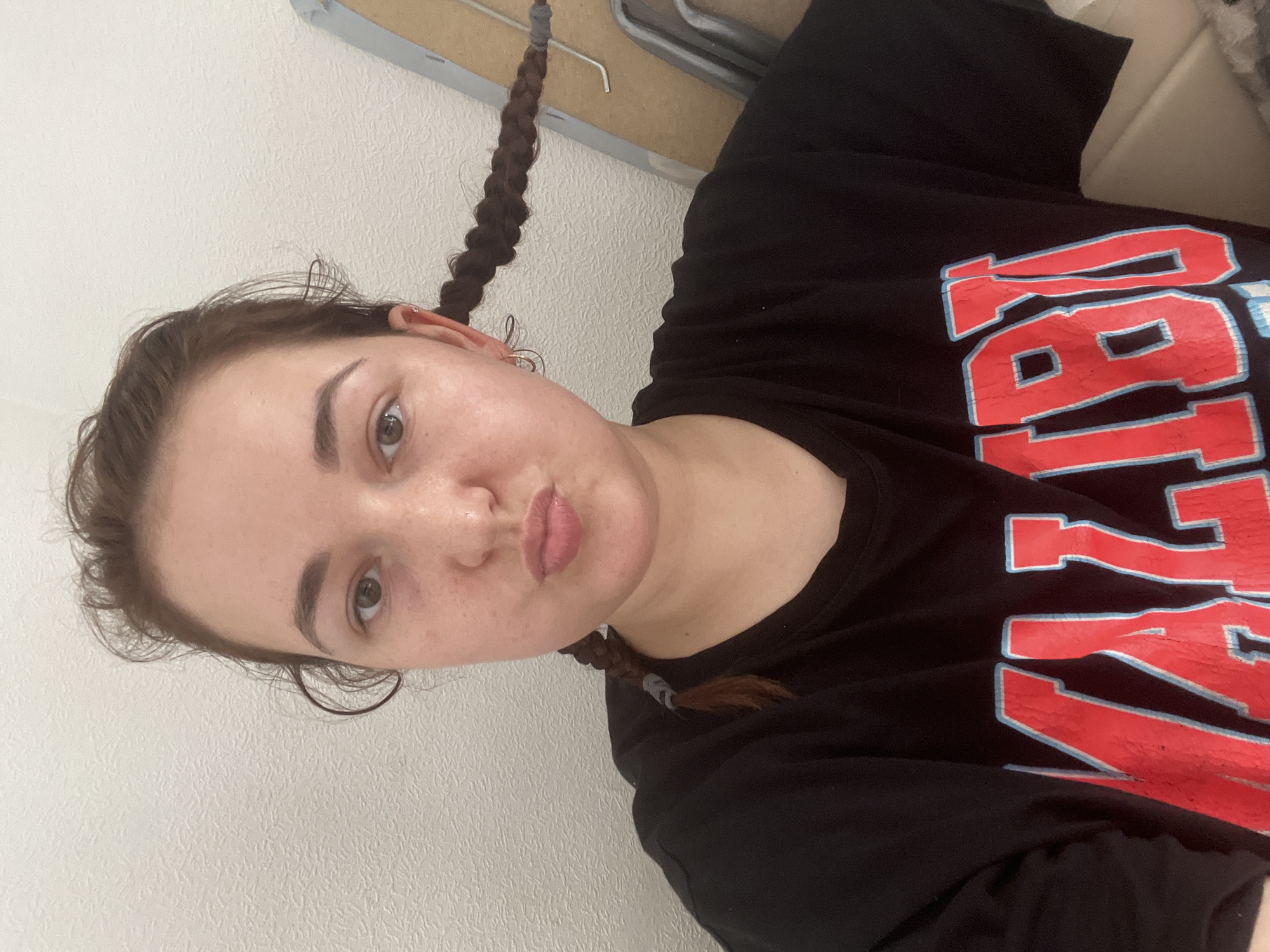A young woman with braided brown hair is taking a selfie in an indoor environment. She is wearing a black t-shirt with red and blue lettering that appears to say "MALIBU". Her hair is styled in two braids, one on each side of her head, and she is puckering her lips. The background appears to be a textured white wall and a light-colored piece of furniture. The image does not contain any information that would allow for the determination of its location in Chelyabinsk, Russia.