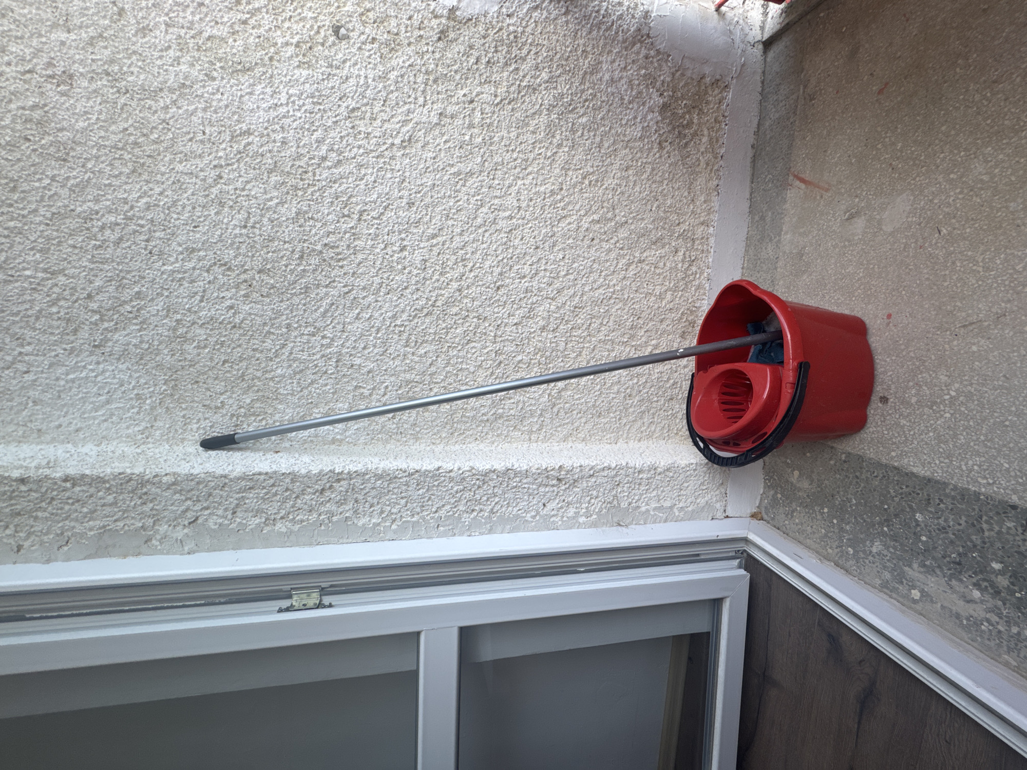 The image captures a utilitarian corner space, likely an indoor or semi-enclosed area such as a utility room or balcony. The main subjects are a red mop bucket with a wringer and a silver metal mop handle, stored against a textured wall.
The wall on the left and spanning the upper center is light-colored, featuring a very rough, granular texture resembling stucco or heavily textured plaster. Adjacent to this, on the right, is another wall of a darker, speckled gray, possibly concrete or a different type of textured plaster. A faint reddish stain or mark is visible on this gray wall. The red mop bucket is prominently displayed, hanging on the right wall in the upper right section of the frame. The mop's silver handle extends from the bucket, resting against the light-colored textured wall.
Across the bottom of the image, a white metal window frame is visible, appearing to be part of a sliding window system with multiple tracks. A small metal hinge or latch is present on the frame. Below the window on the right, a section of dark wooden flooring, possibly laminate with a visible grain pattern, is bordered by a white trim.
The lighting suggests daylight. No human figures are present, and no specific activity is taking place other than the storage of cleaning equipment. There is no visible text in the image. The scene conveys a functional and unadorned environment.