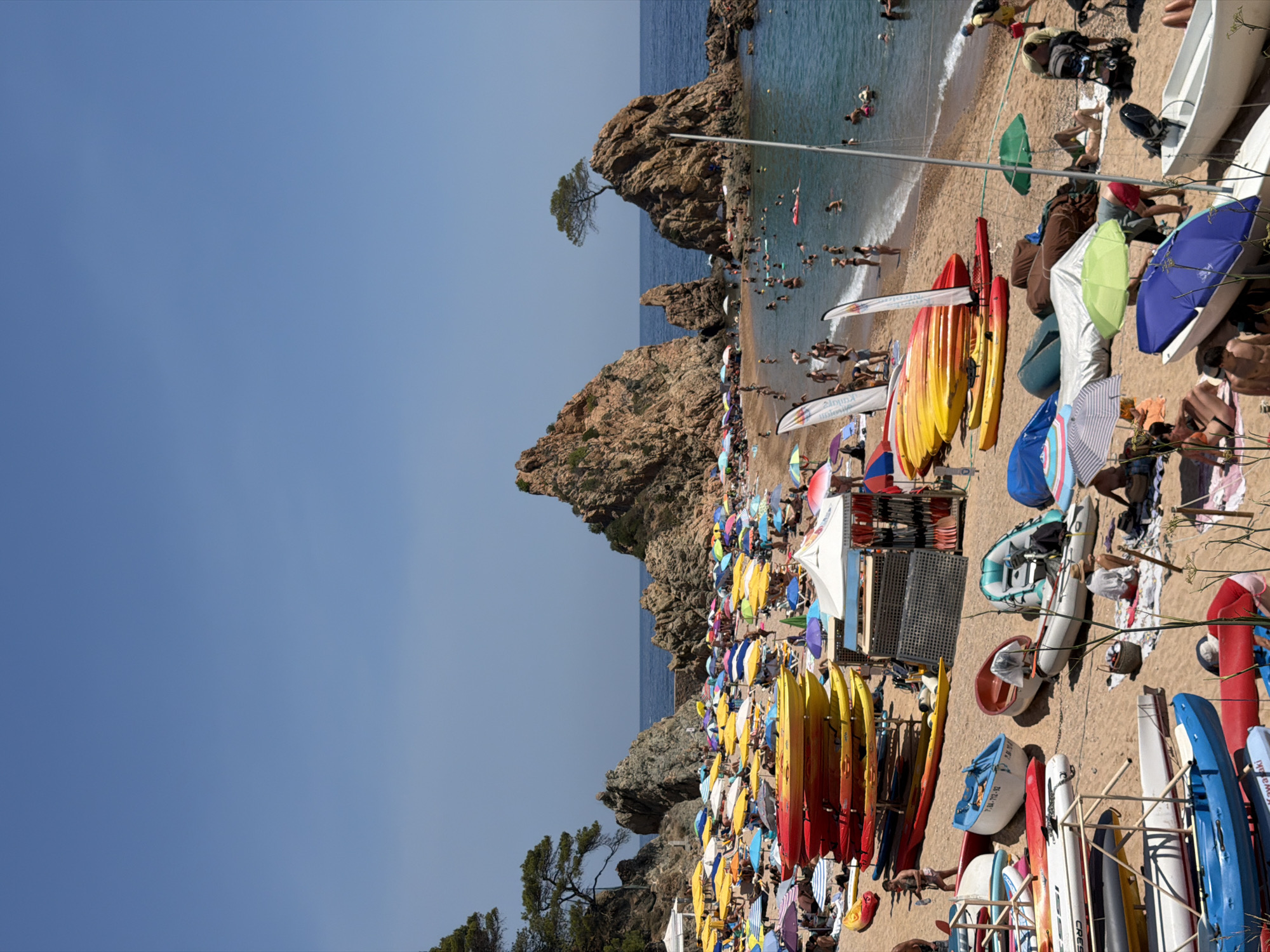 This is a photograph of a crowded beach in Tossa De Mar, Spain. The beach is sandy and slopes down to the sea. Numerous colorful beach umbrellas are spread across the sand, providing shade for beachgoers. A variety of boats, including kayaks and smaller dinghies, are stacked on racks or resting on the sand, suggesting a rental facility or a popular spot for water sports. People are scattered throughout the scene, some sunbathing, others swimming or wading in the clear blue water. The water appears calm with gentle waves. In the background, rocky outcrops rise from the sea, with a single tree growing precariously on one of them. The sky is a clear, bright blue, indicative of a sunny day.