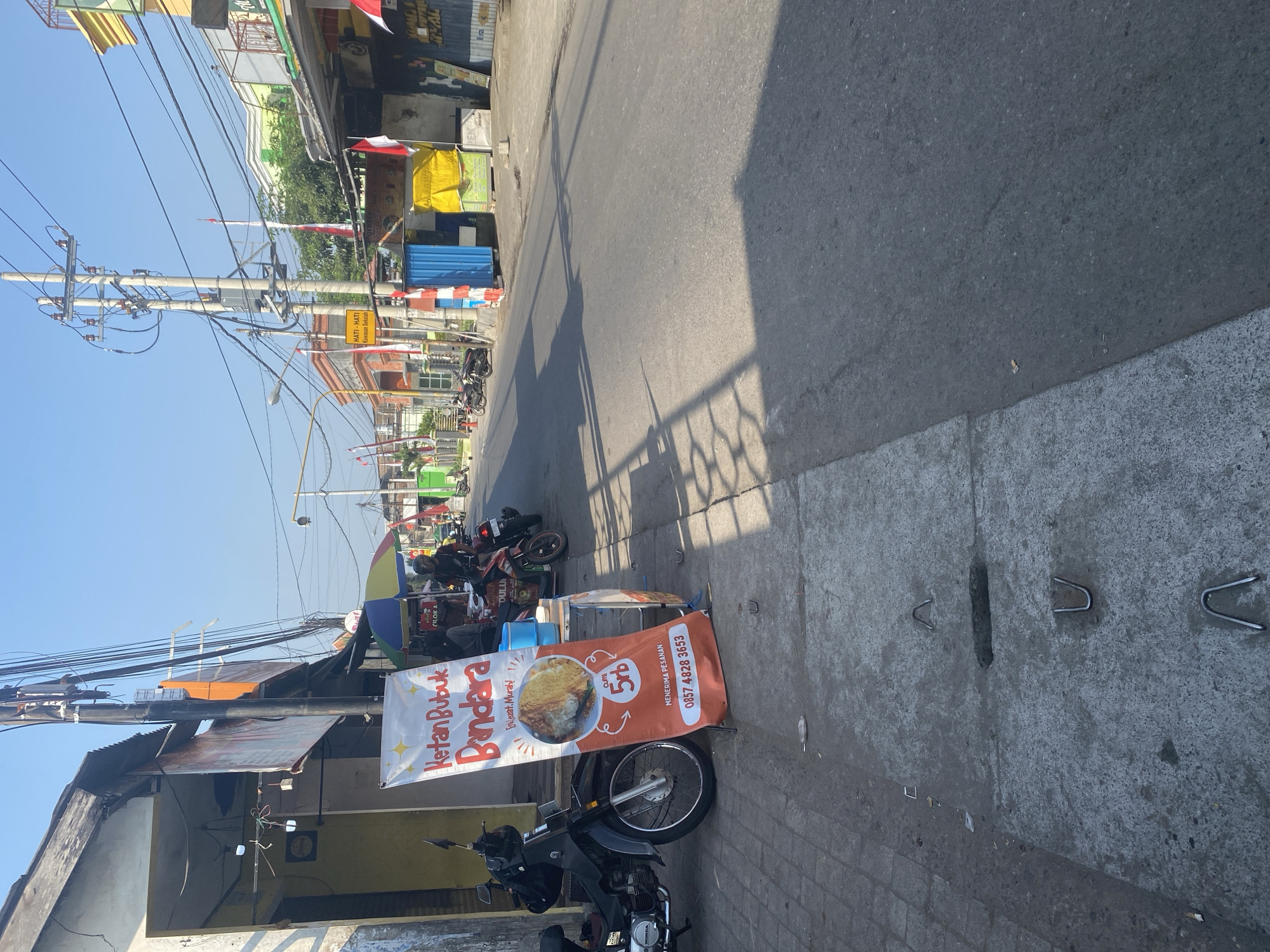 The image shows a street scene in Kediri City, Indonesia, during daytime. A stall selling "Ketan Bubuk Bandara" is set up on the sidewalk, with a prominent orange banner advertising the food, its price (5rb), and a contact number. A motorcycle is parked next to the stall. In the background, several other stalls and buildings line the street, with more motorcycles parked. Indonesian flags are visible on poles, indicating a national celebration or event. The sky is clear and blue. The street is paved, and the sidewalk is made of concrete slabs. There is some litter on the sidewalk. The overall impression is of a bustling street market or commercial area.