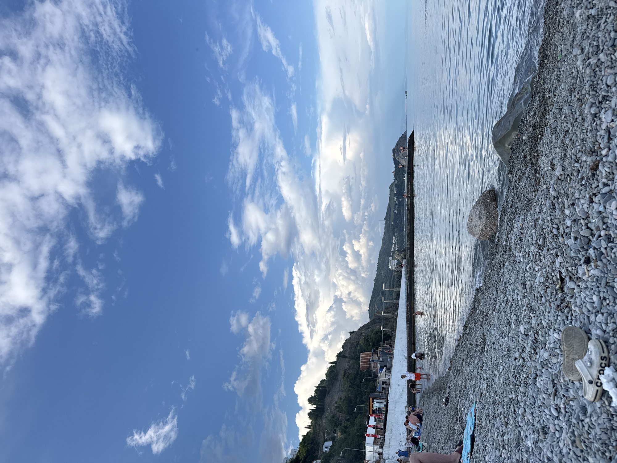 The image depicts a vibrant seaside scene on a sunny day. The sky is a brilliant blue, dotted with fluffy white clouds that stretch across the horizon. A gentle breeze seems to be present, as evidenced by the rippling surface of the calm sea. The shoreline is a picturesque mix of smooth, grey pebbles and a small section of a concrete walkway.
Several people are scattered across the beach and near the water's edge, enjoying the pleasant weather. Some are wading into the shallow water, while others are relaxing on towels or sitting on the pebble beach. The atmosphere appears to be one of leisure and enjoyment, with individuals of various ages participating in typical beach activities.
In the distance, a mountainous landscape rises behind the beach, covered in lush greenery and dotted with buildings. A long pier or breakwater extends into the sea, with a few people visible on it, perhaps fishing or simply enjoying the view. The overall impression is one of a tranquil summer day at a popular coastal destination. There are some faint reflections on the water, suggesting the sun is high in the sky. On the pebble beach, a pair of white Crocs shoes is visible, adding a touch of casual footwear to the scene. The word "CROCS" is clearly legible on the side of the shoe.