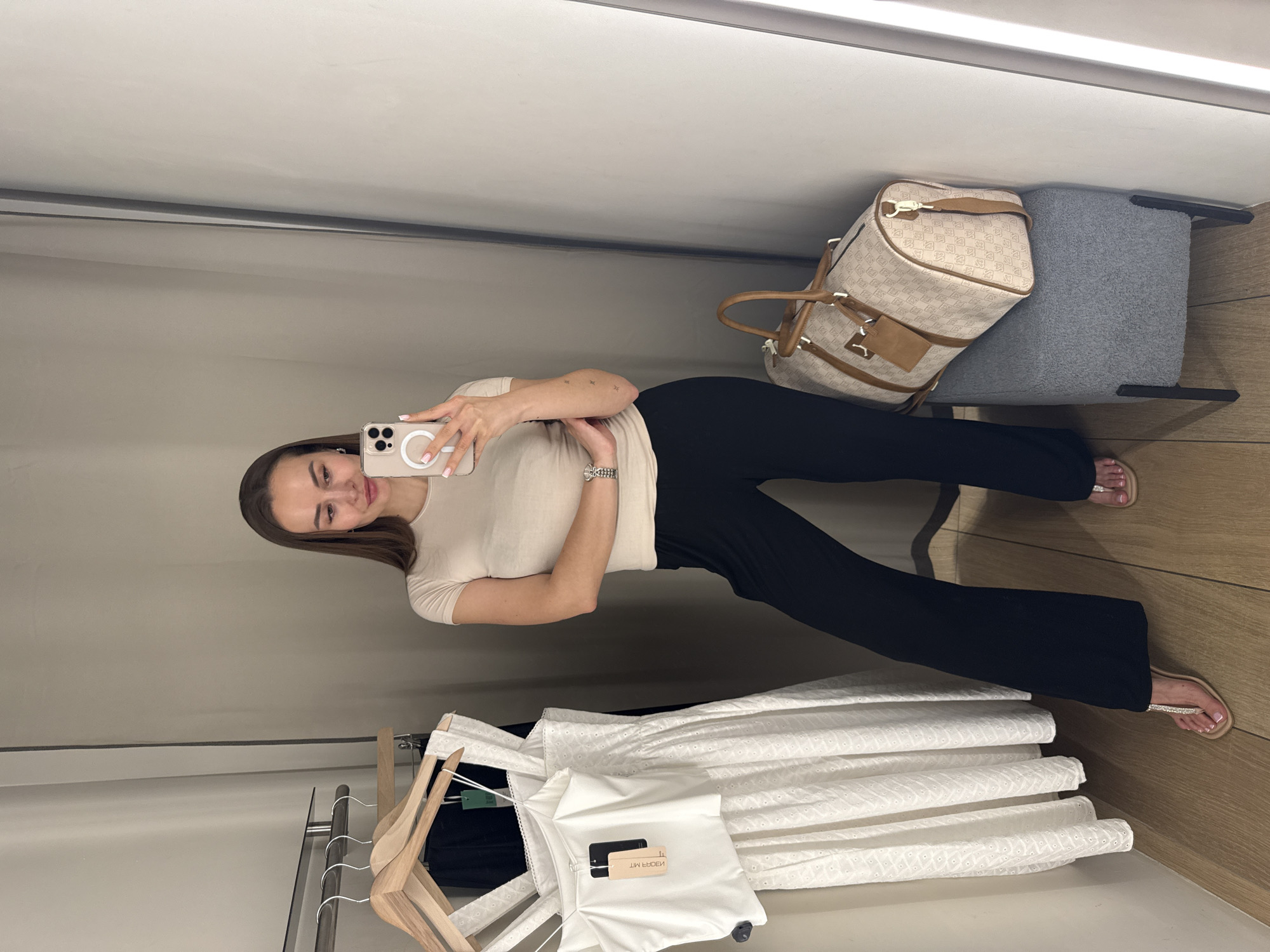 A woman is taking a selfie in a dressing room. She is wearing a beige t-shirt and black pants and holding a smartphone in her right hand. The dressing room has a wooden floor and beige walls. A rack of clothes is visible on the left, with several white dresses hanging on wooden hangers. A beige bag sits on a grey stool in the upper right corner. The dressing room appears to be located in a retail store, likely in Kyiv, Ukraine.