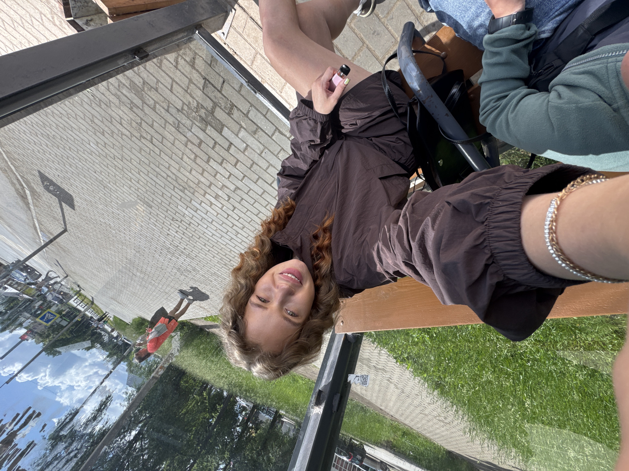 A young woman with curly brown hair is sitting on a wooden bench next to a metal railing, holding a pink vape. She is wearing a dark brown jacket and shorts. In the background, a street in the city of Chaikovsky, Russia is visible, with a paved sidewalk, grass, trees, buildings, and cars. The sky is blue with white clouds. Another person's arm wearing a green sweater and denim jeans is visible in the lower right corner of the image. The image appears to be a selfie, taken from a low angle.