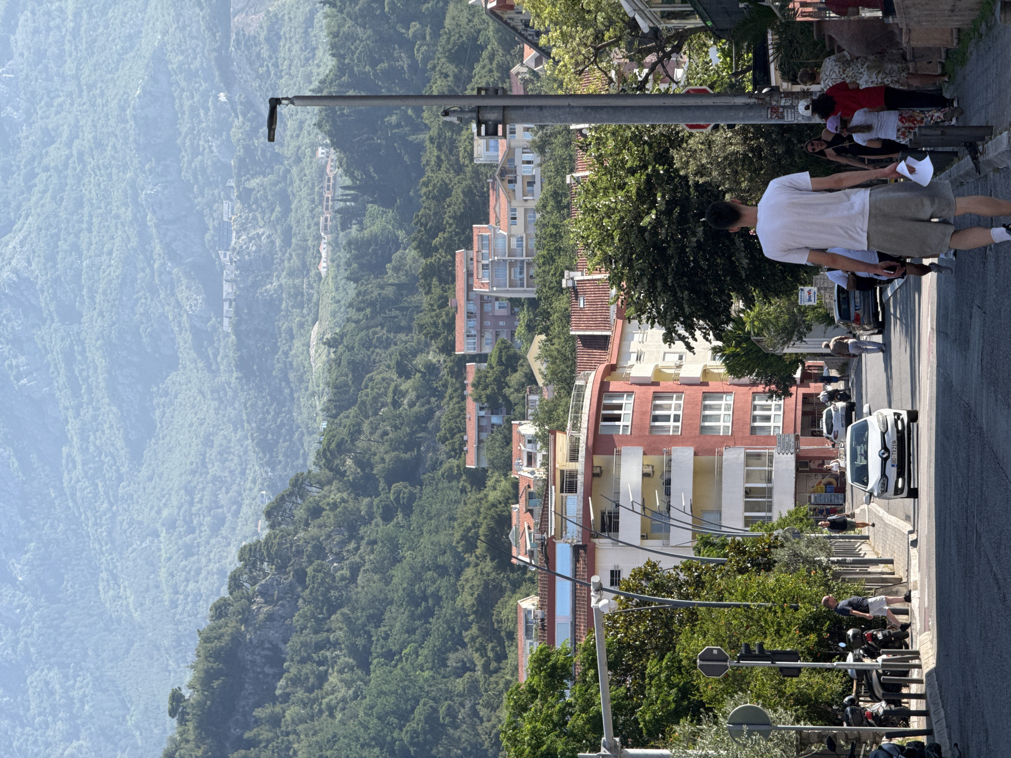 The image shows a street in Budva, Montenegro, with buildings on either side and a steep, tree-covered mountain in the background. The street slopes downwards, and there is a white car driving towards the camera. A man in a white t-shirt and grey shorts is walking away from the camera in the foreground, holding a piece of paper. Several other people are walking on the sidewalk to the right of the street. The buildings visible are a mix of modern apartments and older structures, many with red-tiled roofs. The mountain in the background is densely forested, with some buildings visible on its slopes. The overall scene depicts a sunny day in a coastal town with a mountainous backdrop.