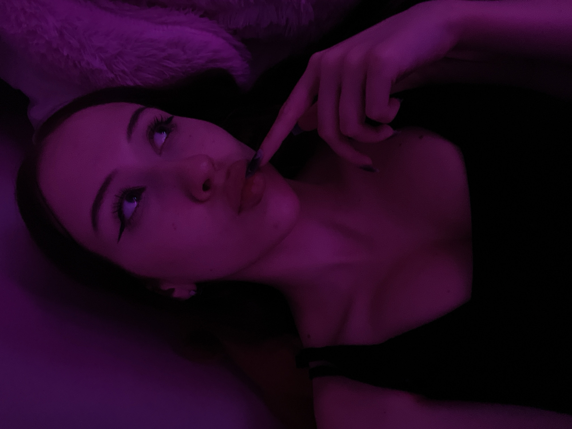 The image is a close-up, low-angle shot of a young woman's face and upper body, illuminated by purple lighting. She is lying down, possibly in bed, with a fluffy textured object visible in the upper right quadrant of the frame. Her eyes are looking upwards and to the right, and she has winged eyeliner and long eyelashes. Her lips are slightly parted, and her index finger is touching her upper lip. She is wearing a black tank top. The overall mood is intimate and moody, with the deep purple lighting casting shadows and highlighting her features. The scene is likely indoors, in a personal setting. There is no visible indication of the city of Votkinsk, Russia.