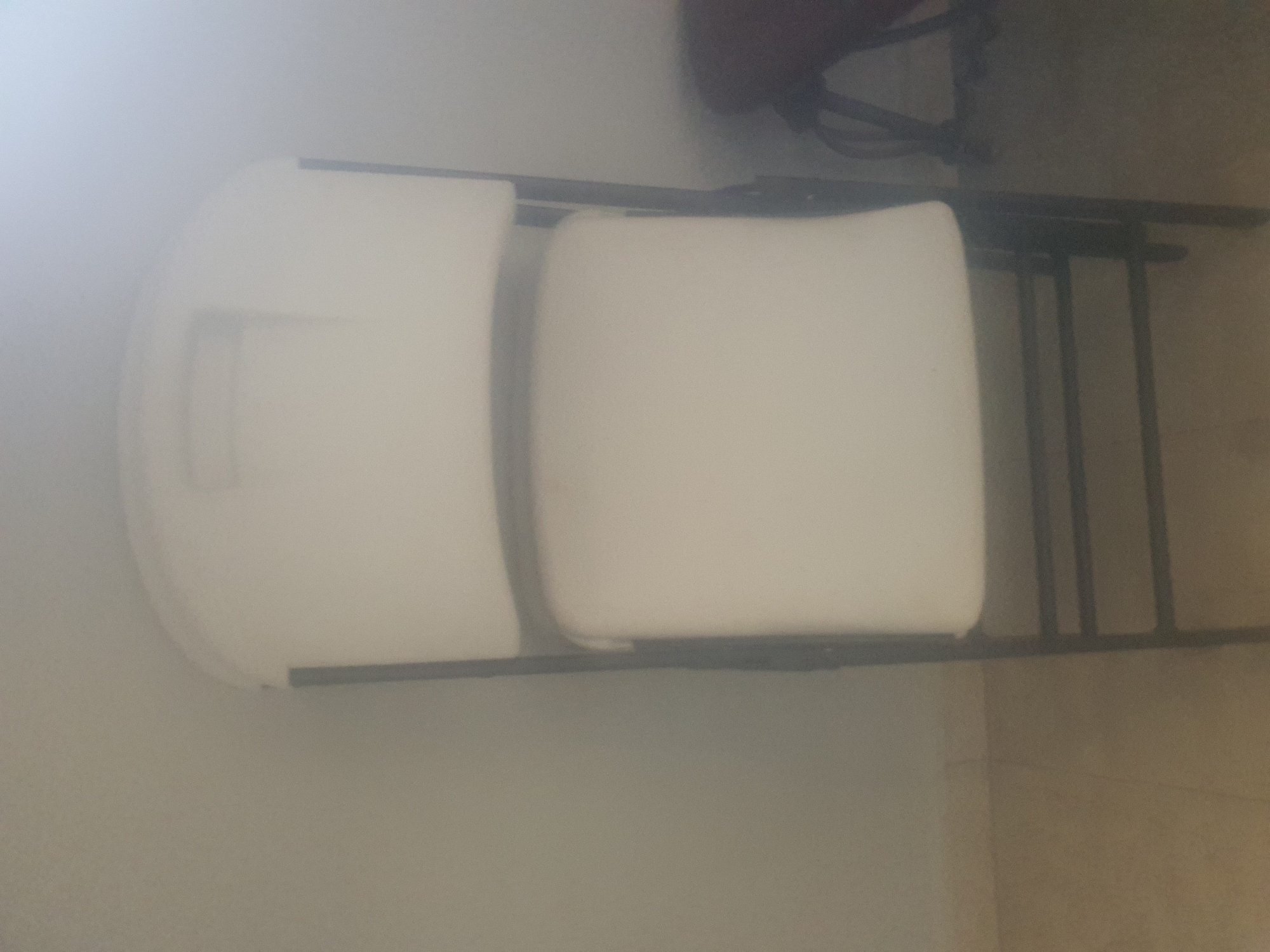 A top-down, slightly angled shot shows a white plastic folding chair positioned in what appears to be an indoor setting. The chair is mostly white, with a black metal frame. The seat and backrest are made of a solid, molded plastic material, with a slightly curved design. The metal frame consists of legs and supporting bars that fold outwards, giving the chair its structure.
The background is muted, with a light-colored wall to the left and a darker, possibly tiled or carpeted floor to the right. The lighting appears to be diffuse, suggesting an interior space with indirect illumination. There is a faint shadow cast by the chair, indicating the direction of the light source.
A dark, indistinct object is visible behind the chair, possibly another chair or a piece of furniture, but it is out of focus and its details are obscured. The overall scene is simple and uncluttered, with the focus primarily on the folding chair. No people are visible in the frame, and the activity taking place is simply the presence of the chair in its setting.
There is no visible text in the image. The time of day and weather are not discernible from the visual information provided. The location context of Jos, Nigeria is not evident from the image itself, as it depicts a generic indoor setting.