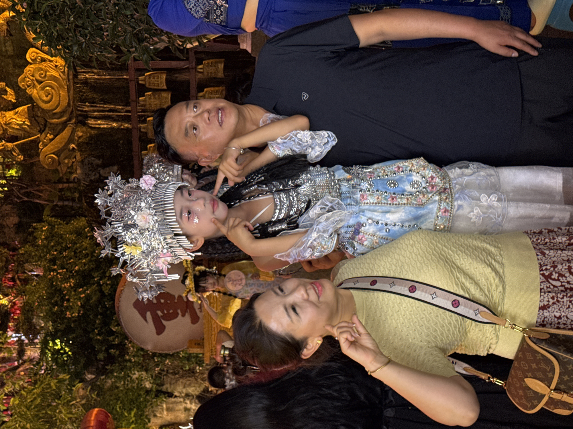 In the city of Yuzhong District, China, a group of people pose for a photograph in what appears to be an outdoor nighttime setting, possibly a cultural festival or performance. The central focus is a young girl dressed in an elaborate, traditional costume with a headdress adorned with silver and pink floral accents. She is also wearing a blue and white garment with intricate beadwork and lace. She touches her cheeks with her fingers, looking towards the camera.
To her right stands a man in a dark blue polo shirt, with his arm around the girl's shoulder. He is looking slightly to the right of the camera.
To the girl's left, a woman with dark hair, wearing a light yellow textured top and a patterned skirt, is also looking towards the camera and touching her own cheek with a finger. She has a brown and tan designer handbag with a patterned strap slung across her body.
In the background, other individuals can be seen, some in costumes. A large red character is visible on a drum. Decorative elements, possibly bells and ornate sculptures, are also present. The lighting suggests a festive atmosphere, with warm lights illuminating the scene and casting shadows. The overall impression is a family or group celebrating a special occasion.