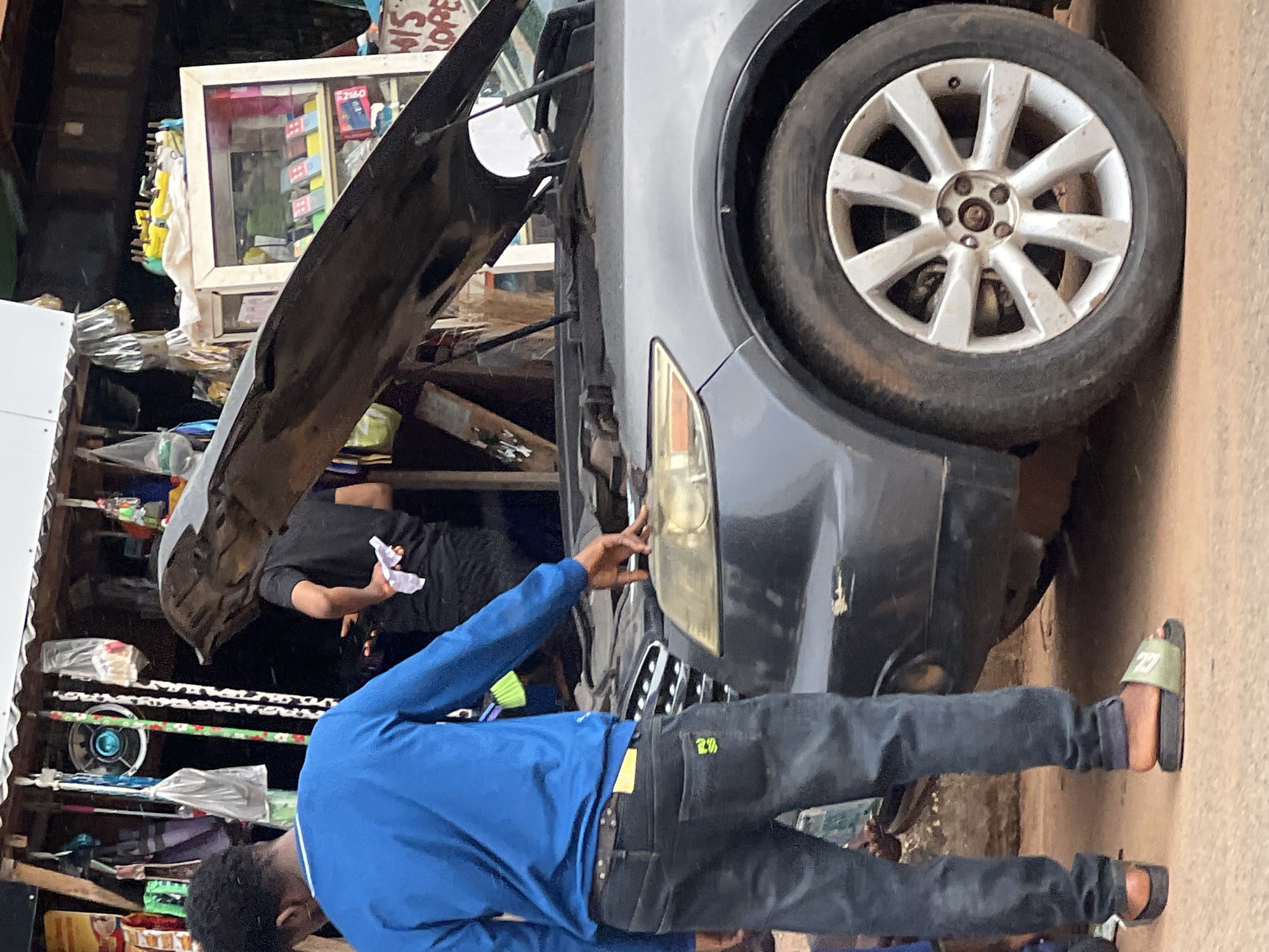 In Okpanam, Nigeria, a dark gray SUV with its hood open is parked in front of a makeshift stall. A young man, wearing a blue long-sleeved shirt and dark jeans, stands with his left hand resting on the vehicle's hood, near the headlight. To his right, another person in a black shirt stands with their back partially turned, holding a white cloth. The stall behind them displays various items, including what appear to be cleaning supplies, bags, and a small refrigerated display case filled with packaged goods. The ground is a mix of dirt and pavement.