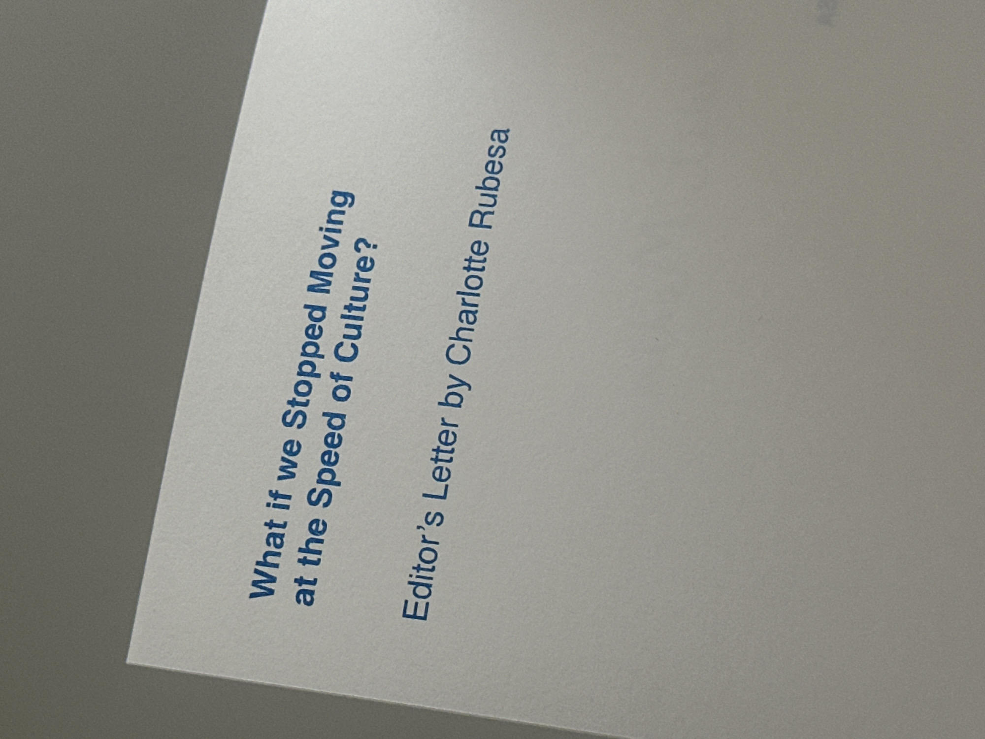 The image shows a piece of paper with text printed in blue ink. The text reads "What if we Stopped Moving at the Speed of Culture?" followed by "Editor's Letter by Charlotte Rubesa." The paper is placed against a gray background. The image does not provide information about a location in Tunbridge Wells, United Kingdom, people, objects, actions, interactions, or the environment.