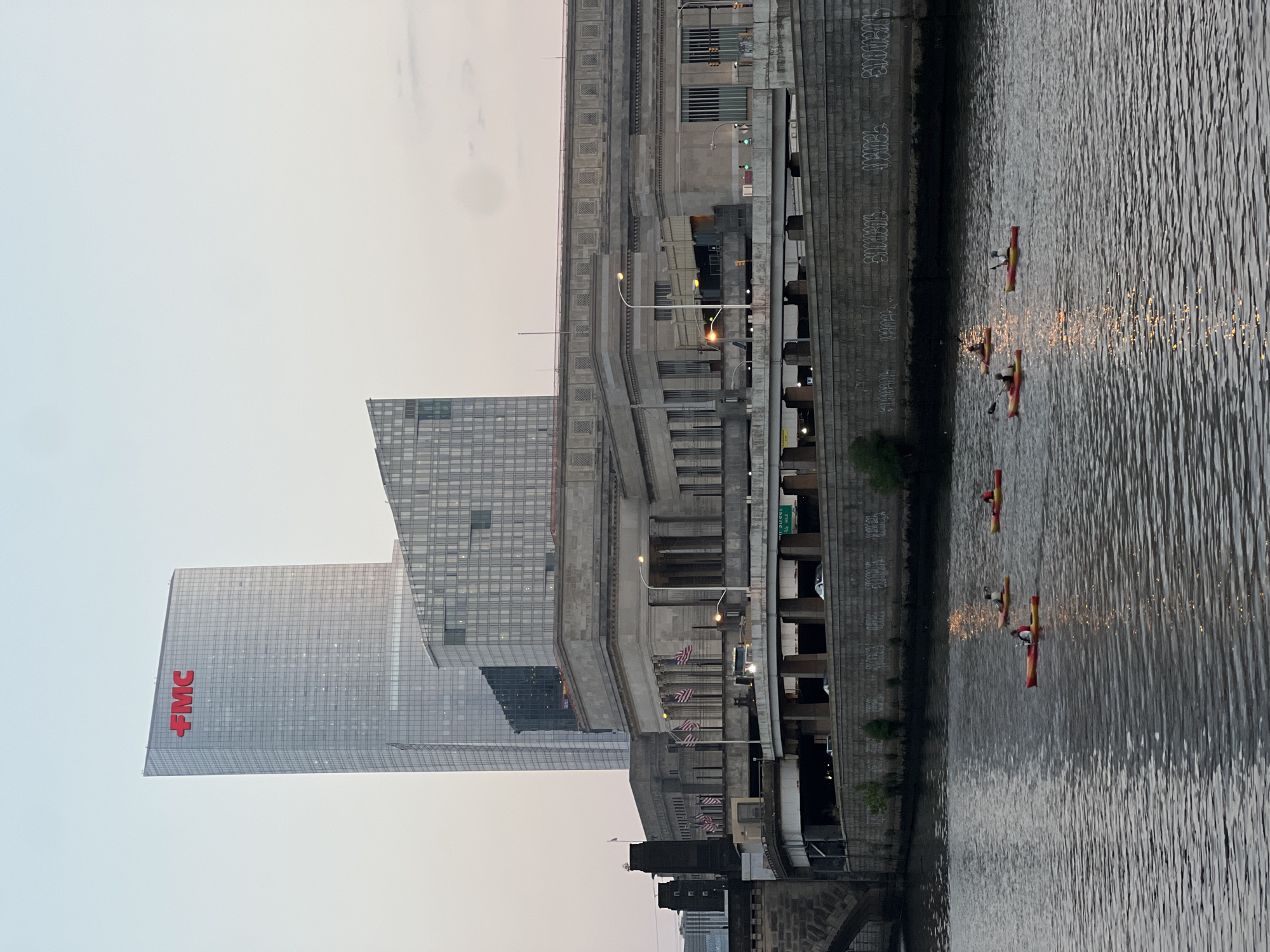 The image shows a view of the Delaware River in Philadelphia, Pennsylvania, USA. In the background, there is a tall, modern glass skyscraper with the red "FMC" logo at the top, likely FMC Corporation headquarters. Next to it, a shorter building with a glass facade and geometric design is visible. In front of these modern structures, a large, classical-style stone building with columns and rows of windows stands, likely a historic train station. A highway overpass runs in front of the stone building, with cars visible on it. The river in the foreground has several people in kayaks, paddling on the water. The sky is overcast with a soft, diffused light, suggesting either early morning or late evening. Graffiti is visible on the concrete wall along the riverbank.
