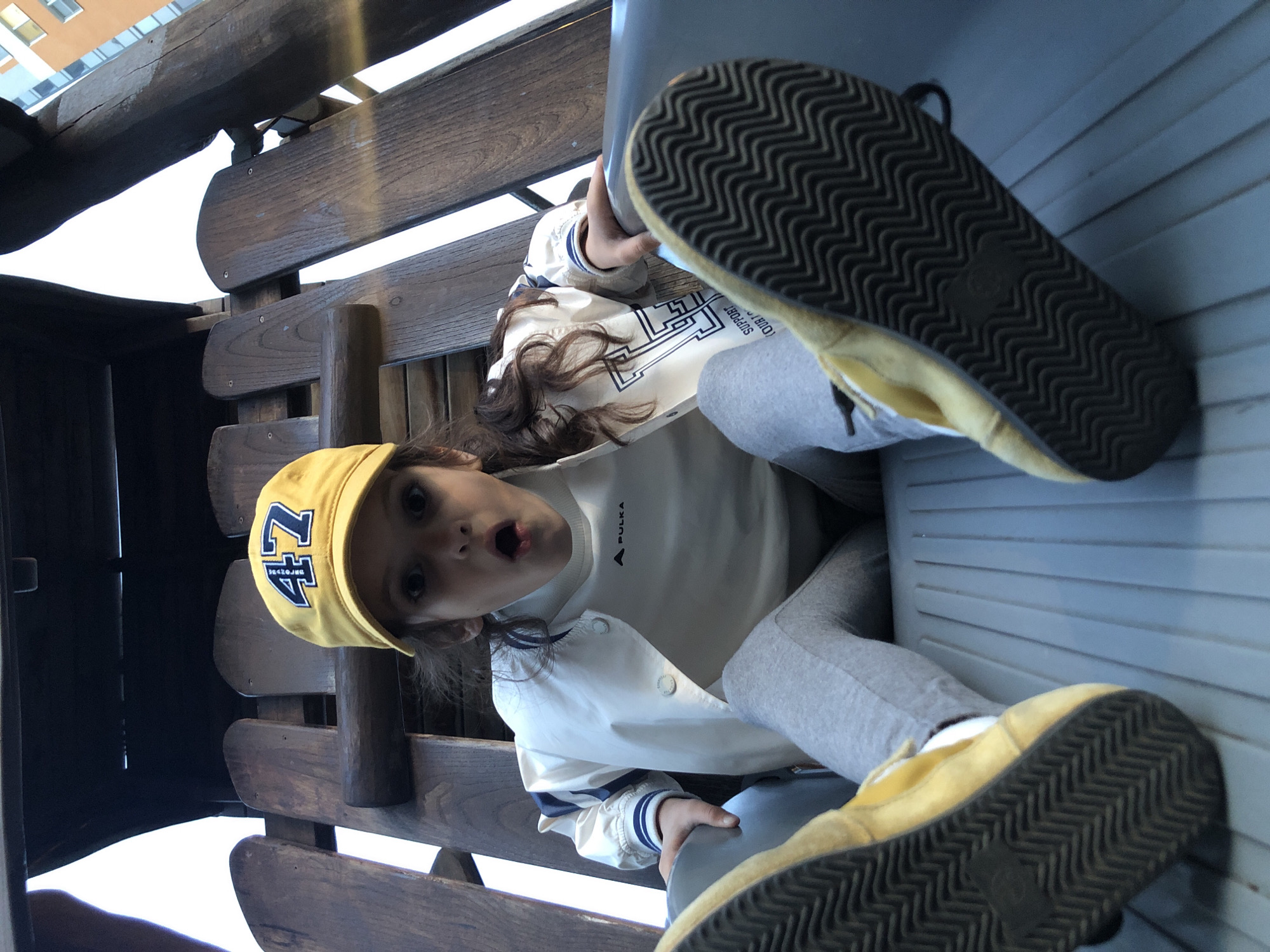 This image, rotated 90 degrees clockwise, captures a young child with dark hair looking directly at the camera with an open-mouthed, surprised, or playful expression. The child is wearing a bright yellow baseball cap featuring the number "47" in dark blue.
She is dressed in a white long-sleeved shirt with the brand "PULKA" and a small triangular logo visible on the chest. Over this, she wears a light-colored (possibly off-white) bomber jacket with blue striped cuffs and large, circular white buttons. A complex graphic design is on the jacket, with partially visible text that includes "SUPPORT YOUR...". The child is also wearing grey sweatpants and bright yellow slip-on shoes with prominent dark soles featuring a wavy, zig-zag tread pattern. One shoe is in the foreground, with its sole facing the viewer.
The child is seated on a sturdy wooden structure, possibly a bench or swing, made of thick planks and beams. Her legs are propped up against a light grey, ridged surface, which could be a wall or a part of another structure. Through gaps in the wooden seating, parts of a distant building with orange and light blue sections are visible against a bright background, indicating it is daytime. The location context provided is Saint Petersburg, Russia.