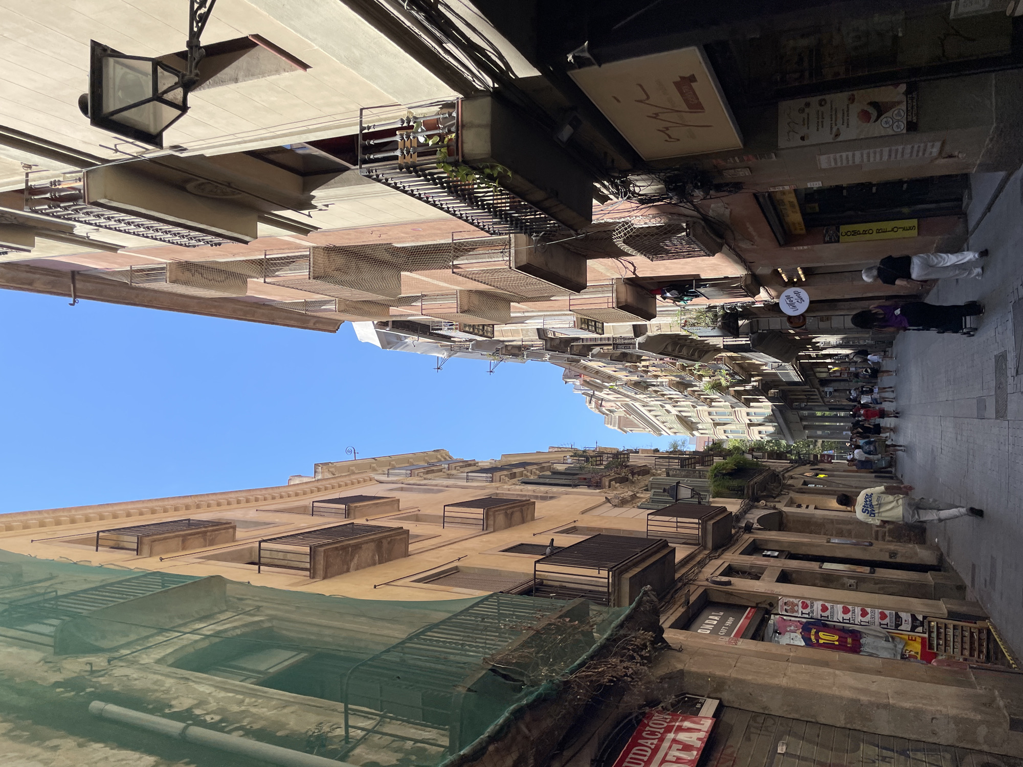 This is a vertical, eye-level shot of a narrow street in Barcelona, Spain, lined with tall, multi-story buildings. The architecture features balconies with ornate railings, some adorned with plants. One building on the left is partially covered by green netting, suggesting ongoing renovation. Shops line the ground floor of the buildings, with various signs and displays visible, including a "LIQUIDACION TOTAL" sign and posters for what appears to be a football player. Several people are walking down the street, with a man in a light yellow t-shirt walking away from the camera and two women with a stroller walking towards the camera. The sky above is clear and blue. The overall impression is of a bustling, historic urban environment on a sunny day.