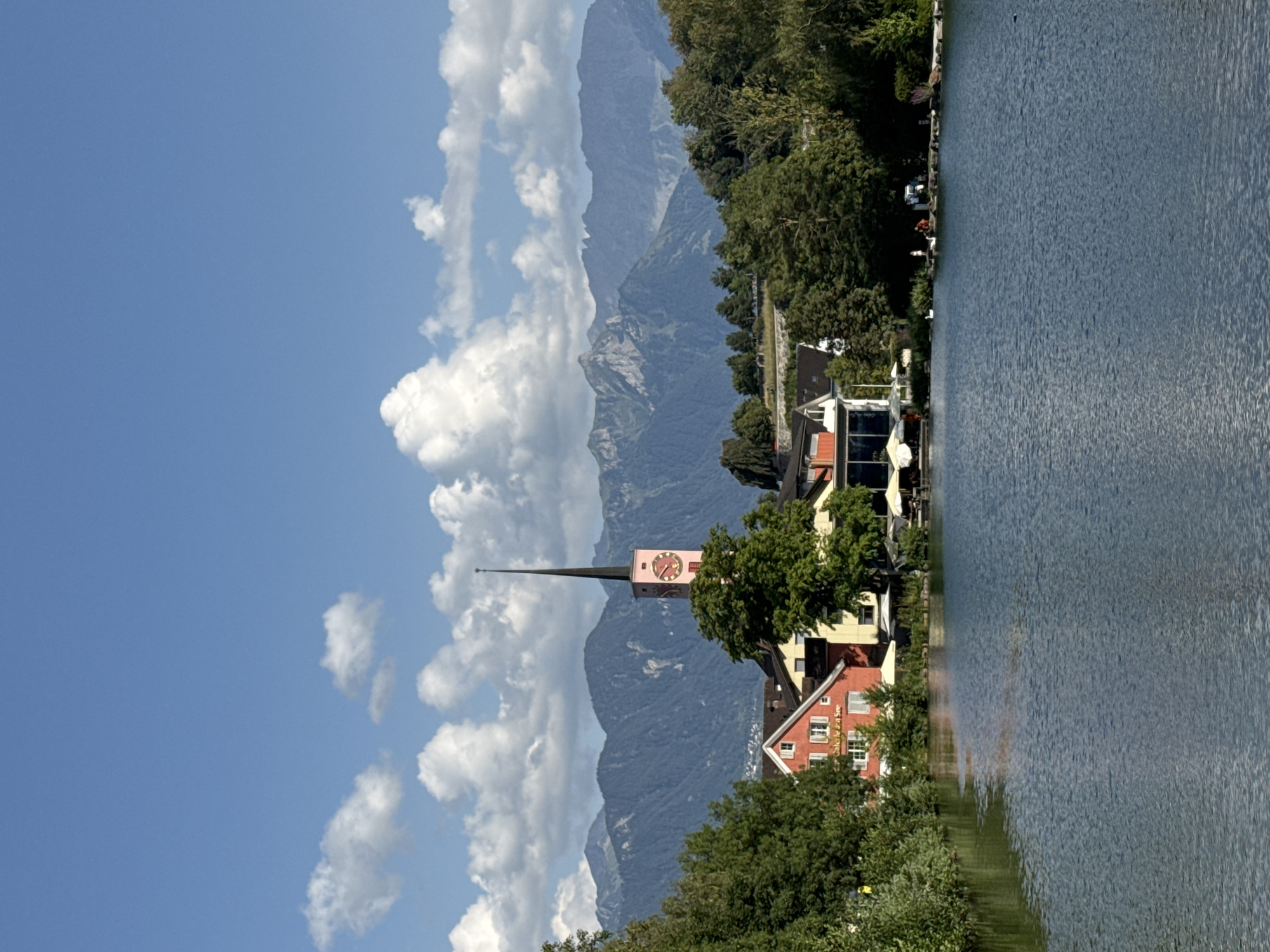 This is a photograph of a scenic lake in Buchs (SG), Switzerland. In the foreground, a calm body of blue water reflects the sky and surrounding scenery. On the far bank of the lake, a cluster of buildings is visible. One prominent building is red with white trim and windows. Next to it, a yellow building can be seen. A tall, slender clock tower with a pink face and a dark spire rises above the buildings, its hands indicating the time. Behind the buildings, a dense line of green trees creates a natural barrier. Further in the background, a range of imposing, blue-tinted mountains stretches across the horizon, their peaks partially obscured by the haze of distance. The sky above is a bright blue, dotted with fluffy white clouds. Along the edge of the lake, several white umbrellas are set up, suggesting an outdoor seating area for a cafe or restaurant. A few individuals are present near the water's edge, with two white swans also visible.