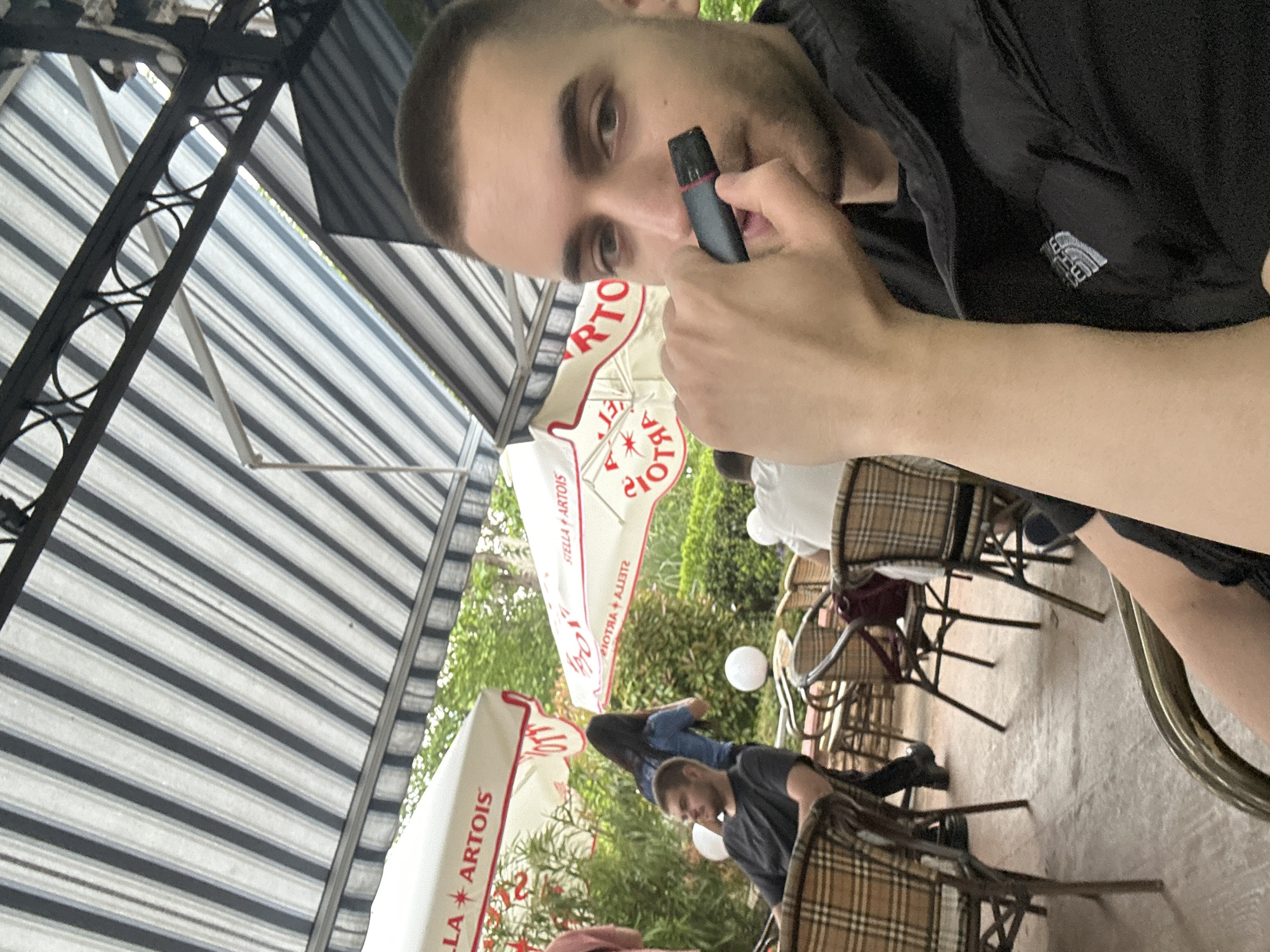 A man with short hair is holding a vaping device up to his mouth. He is wearing a black shirt with The North Face logo on his left chest, and sitting in a chair. The environment appears to be an outdoor seating area, with Stella Artois branded umbrellas visible. Other patrons are present, also seated at tables. The location appears to be a patio or similar outdoor dining space. Based on the Stella Artois umbrellas, it is most likely in a commercial district, but without additional information, the specific location in Burgas is impossible to discern. The image appears to have been taken with a cell phone.
