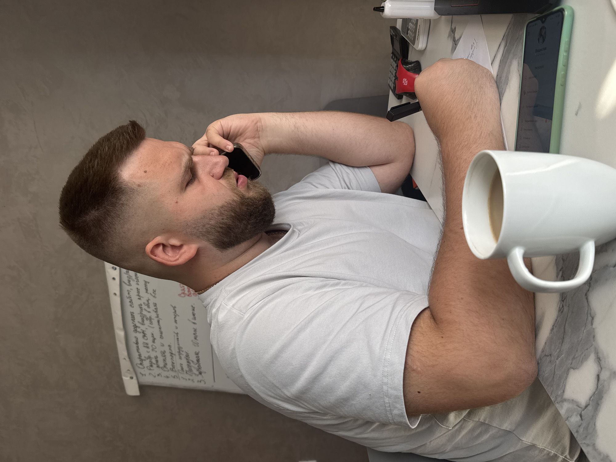 The image shows a man with a beard and a short haircut sitting at a table, talking on a mobile phone. He is wearing a white t-shirt and his left arm is resting on the table. In front of him, there is a white mug with coffee and a smartphone. There are also a calculator and a pen on the table. In the background, there is a whiteboard with some writing on it. The overall scene appears to be an office or home office environment. The image does not provide any specific geographical indicators to suggest a location in Sochi, Russia.