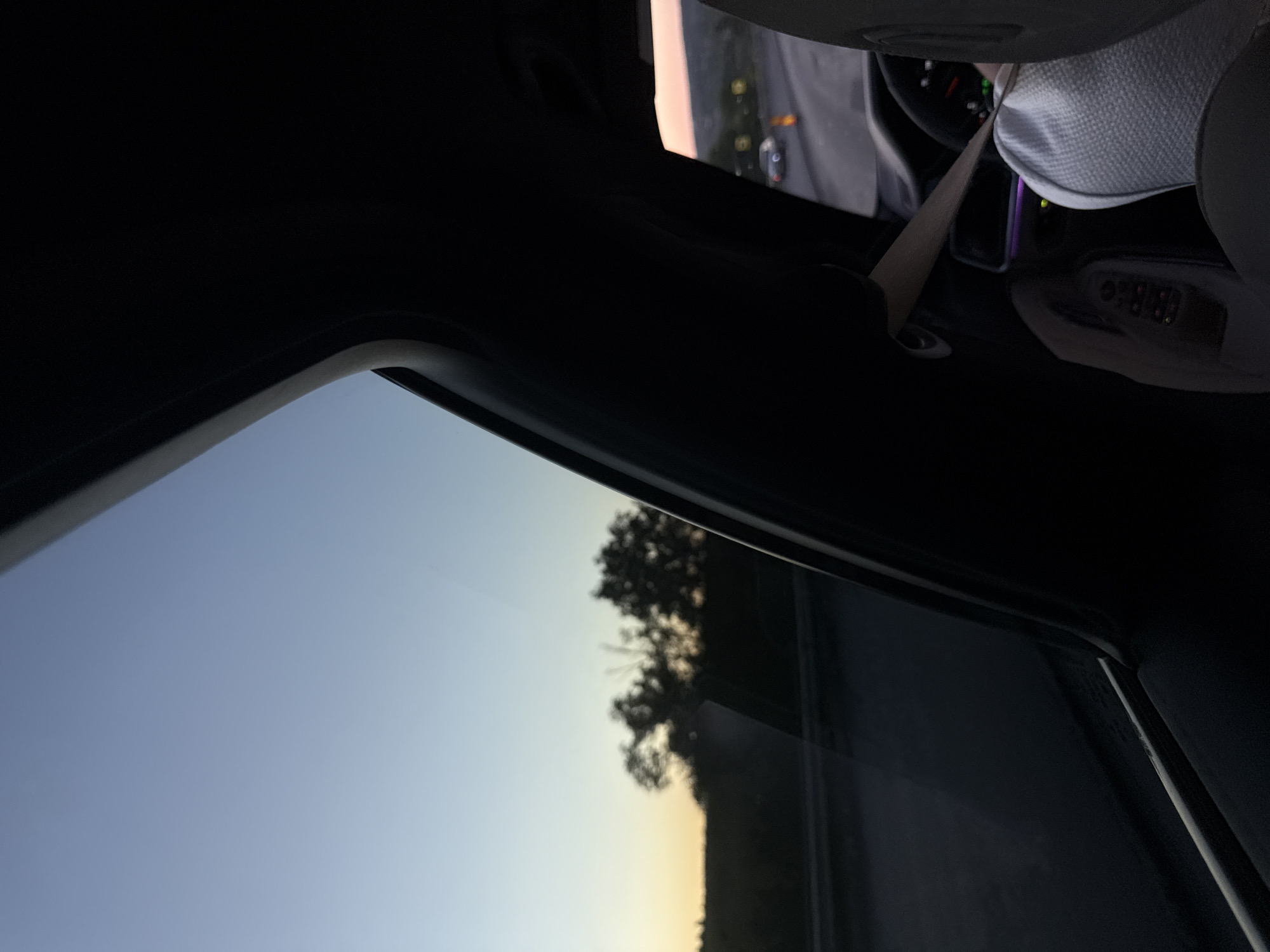 The image depicts a view from inside a car at dusk. The sky is a gradient of blue to pale orange near the horizon. Silhouetted trees are visible against the sky on the left side of the frame, along with a road and what appears to be a highway. Inside the car, the dashboard and a seat are visible. A person's feet, wearing white patterned socks, are visible in the lower right portion of the frame, resting on the car floor. The ambient lighting inside the car shows purple and green accents. The scene is consistent with a person traveling by car on a road near Fundul Galbenei, Moldova, during twilight.
