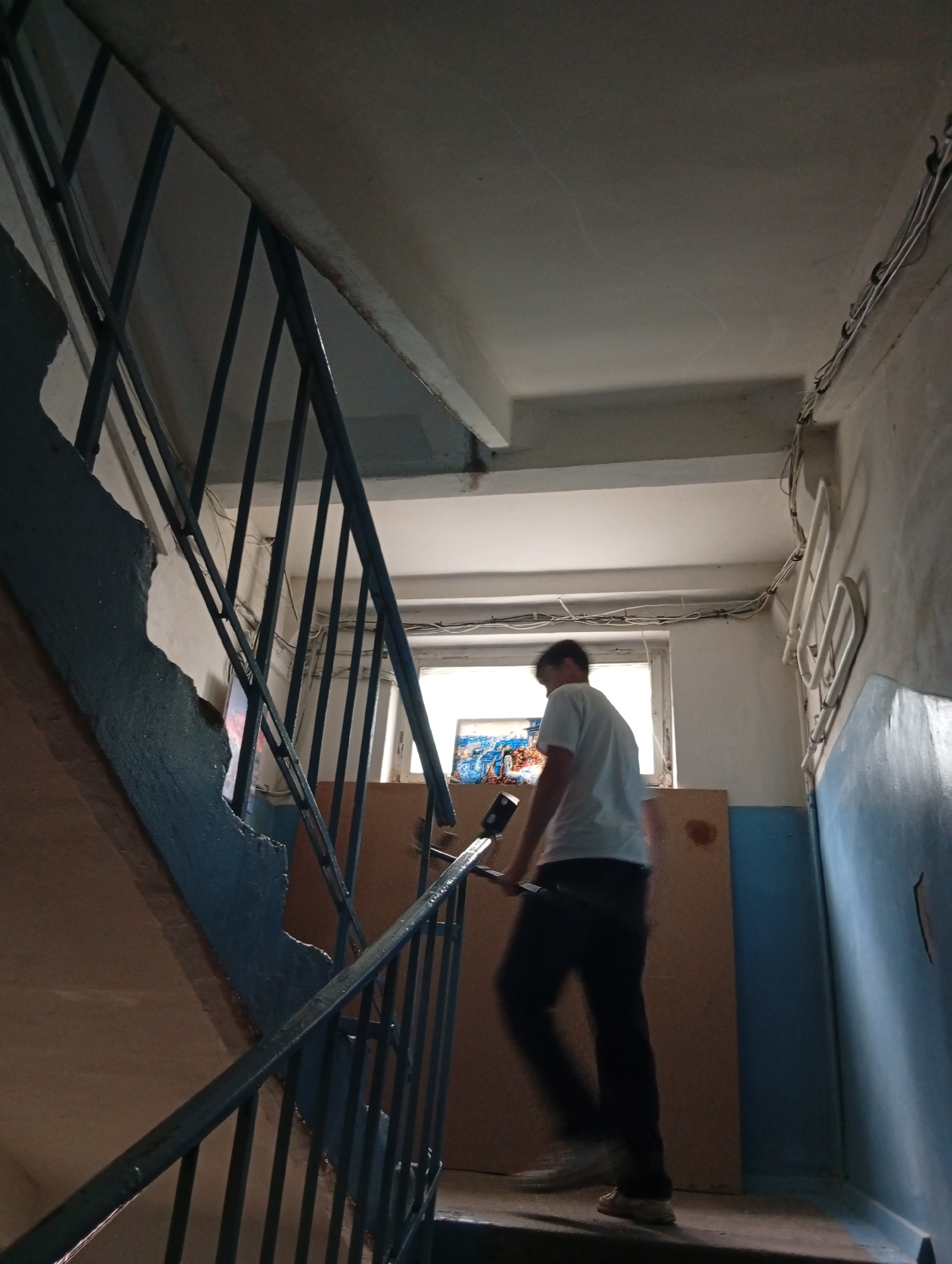 The image captures an indoor stairwell in what appears to be an older building in Poltava, Ukraine. The scene is illuminated by natural light streaming through a rectangular window at a landing, indicating daytime.
A person, likely a young male, is depicted mid-stride, ascending the stairs. He is viewed from a side-rear angle, wearing a white t-shirt, dark trousers, and light-colored shoes. His figure is slightly blurred, suggesting movement, and he holds an unidentified elongated object in his right hand.
The staircase is made of concrete with a visible, worn, and chipped blue-painted edge. A dark metal railing with vertical bars runs alongside the stairs. The walls show signs of age, with sections painted in faded light blue and off-white. Exposed electrical wiring is visible, running along the ceiling and walls, secured by clips. To the right of the window, a white, multi-barred heating fixture or radiator is mounted on the wall. Below the window, several wooden or cardboard panels are propped up. Through the window, a colorful, indistinct object can be vaguely seen.
The environment conveys a utilitarian and somewhat unkempt atmosphere, typical of common areas in older residential or public buildings. There is no discernible text visible in the image, and the overall mood is neutral, without strong emotional cues.