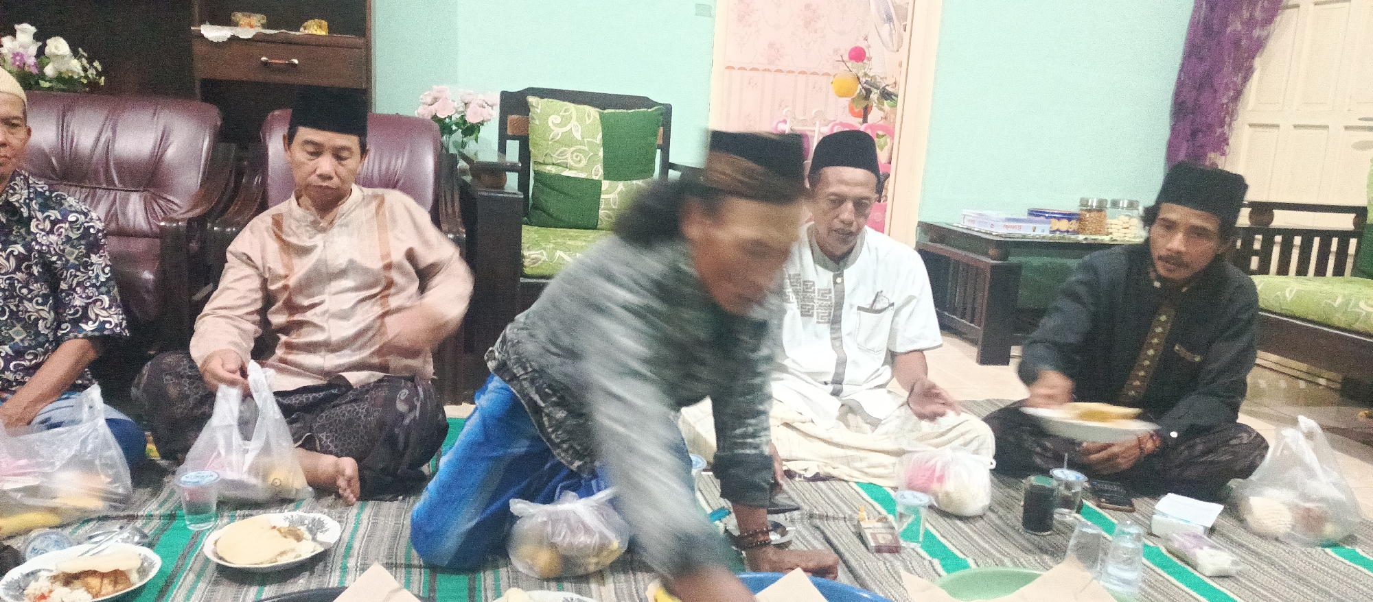 The image depicts an indoor gathering of at least five Indonesian men, likely in the city of Arjasa, Indonesia, judging by the context of other images from this set. The men are seated on a striped mat on the floor, which appears to be a living room or common area within a home. The lighting suggests it is evening or a dimly lit indoor space.
In the foreground, a man in a grey jacket and blue pants is blurred as he leans forward, possibly reaching for food or interacting with someone out of frame. To his left, a man wearing a patterned shirt and a peci (traditional cap) sits with a plastic bag of food in his lap. To his right, a man in a light brown, patterned garment and a peci is also seated, holding a plastic bag.
Further back, a man in a white shirt with a patterned neckline and a peci is seated with his hands clasped. To his right, a man in a dark patterned outfit and a peci is holding a white plate with food.
The environment includes furniture like plush chairs and a wooden cabinet. Decorative items such as flowers and cushions are visible. Food items, plates, plastic bags, glasses, and a cigarette pack are scattered on the mat, indicating a meal or gathering is in progress. The overall impression is one of a communal event, possibly a religious or family gathering, given the attire and cultural context suggested by the peci caps.