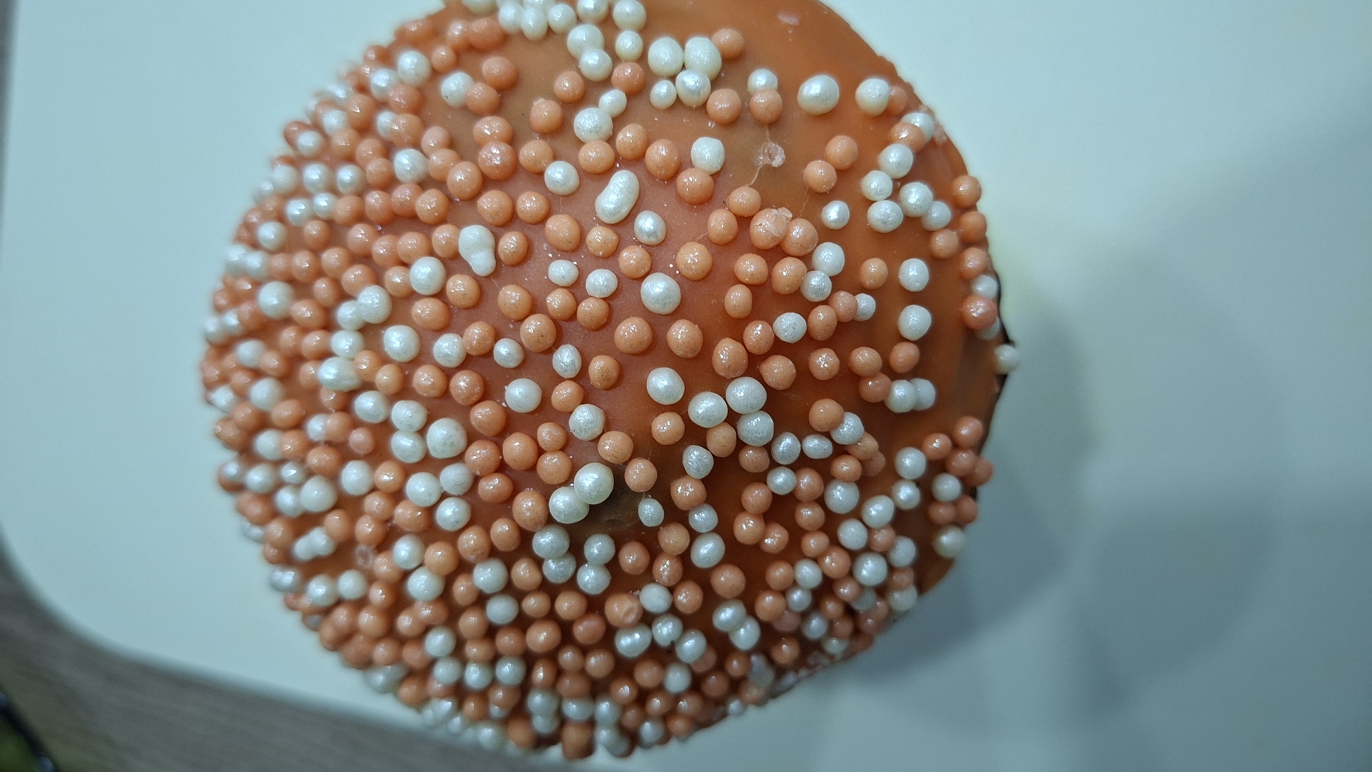 A close-up overhead shot reveals a spherical dessert, potentially a cupcake or cake ball, coated in orange icing and densely covered with small, spherical sugar sprinkles in alternating orange and white colors. The dessert rests on a light-colored surface, likely a plate or tray, with hints of a wooden edge and possibly another object partially visible in the lower left corner. The location of Chaikovsky, Russia is not discernible from the image.