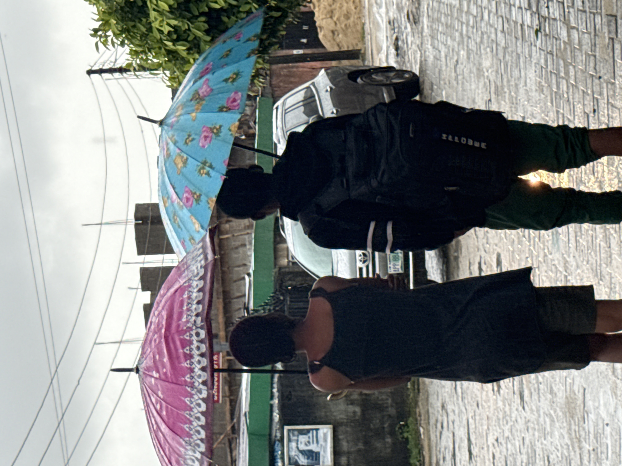 Two individuals, viewed from behind, are walking under umbrellas on a paved street in Ado, Nigeria. The person on the left is holding a pink umbrella with a decorative border and wearing a dark tank top and shorts. The person on the right is carrying a black backpack and is under a light blue umbrella adorned with pink and yellow flowers. They are walking away from the viewer on a wet, brick-paved surface, suggesting recent rain. In the background, a silver SUV is parked, and there are buildings and foliage visible. The sky is overcast, contributing to the dim lighting of the scene.