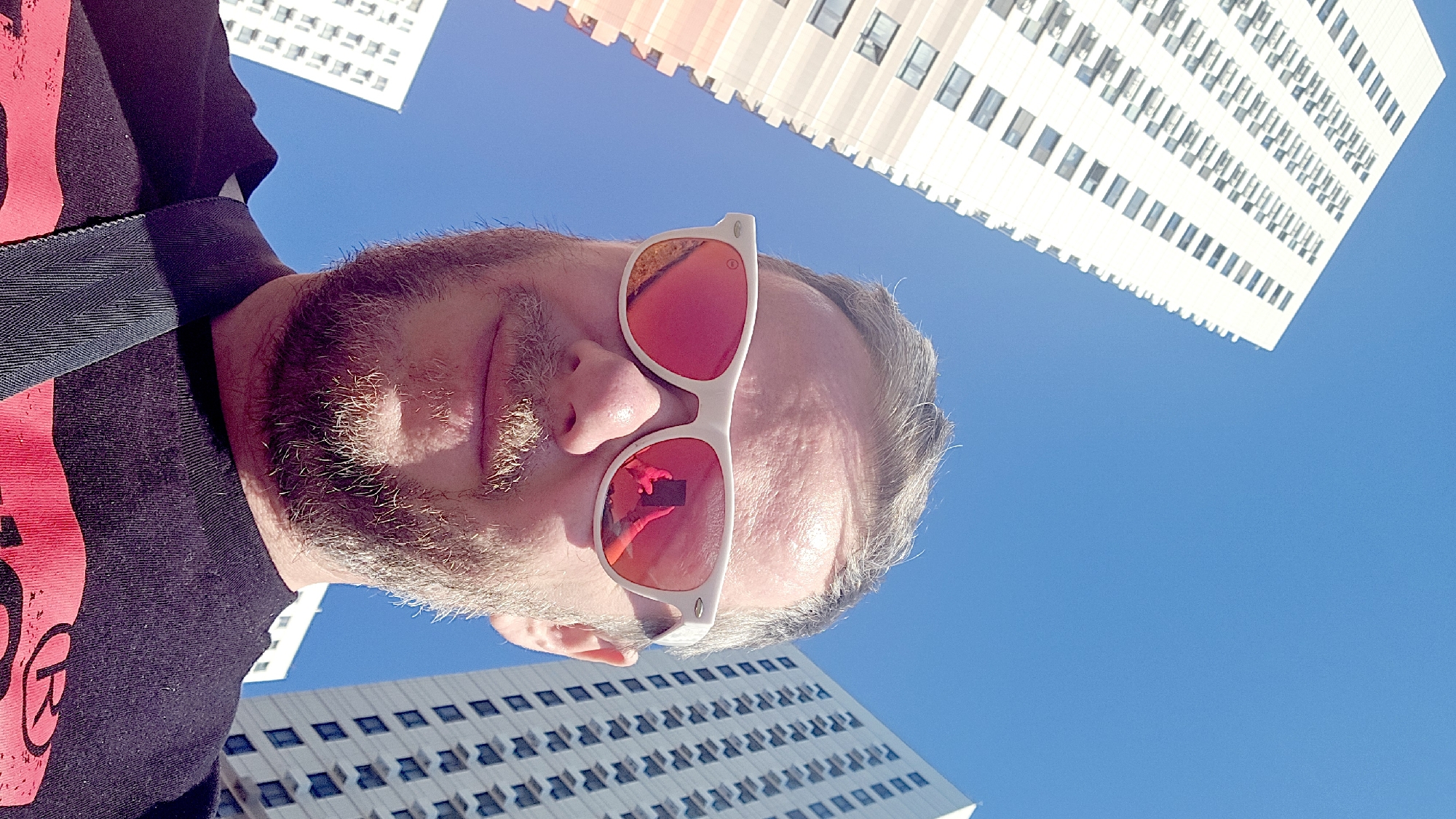 A man wearing white-rimmed sunglasses with reflective lenses is photographed from a low angle, framing his face against a clear blue sky and several white high-rise buildings. The buildings are characterized by a repetitive pattern of windows. The man has a beard and is wearing a dark t-shirt with a red graphic on the shoulder, and a strap is slung across his shoulder. The location is in Moscow, Russia.