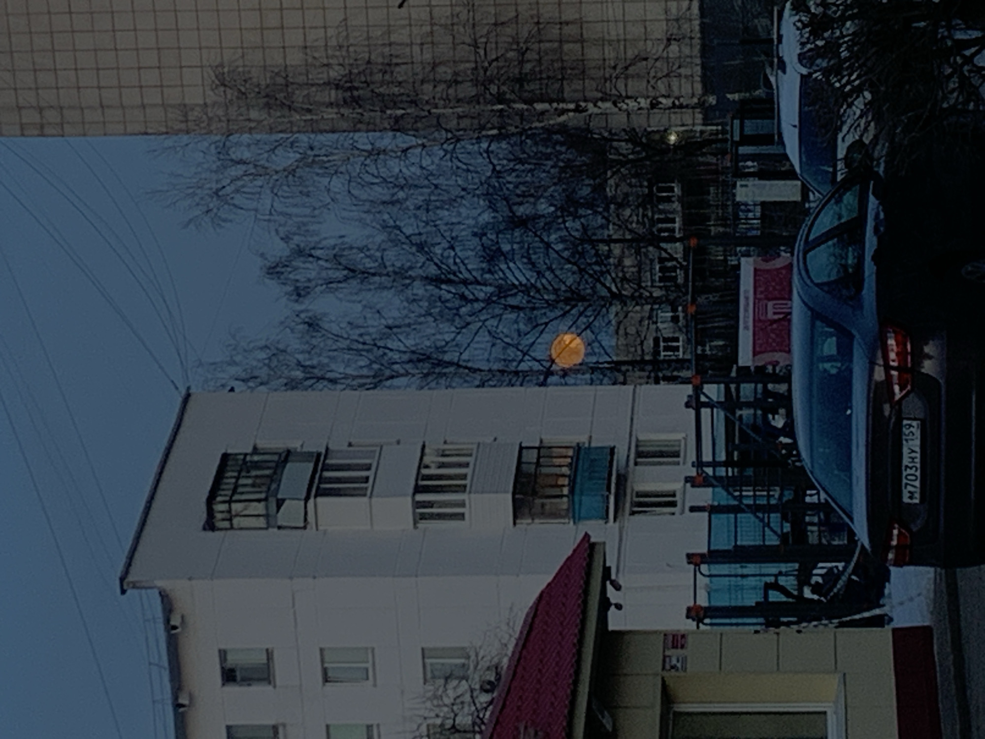 The image shows a street scene in Chaikovsky, Russia, with a multi-story white building dominating the view. The building has numerous windows, some of which have small balconies. A red roof is partially visible in front of the building. In the upper portion of the image, bare tree branches spread across the scene, partially obscuring the sky. A bright, circular orange object resembling the moon or a street lamp is visible through the branches. A dark-colored car is parked on the right side of the frame, with its license plate partially visible as "M703HY 159". The environment seems urban, with another building appearing in the background, slightly out of focus. The sky is a muted blue, suggesting either early morning or late evening.