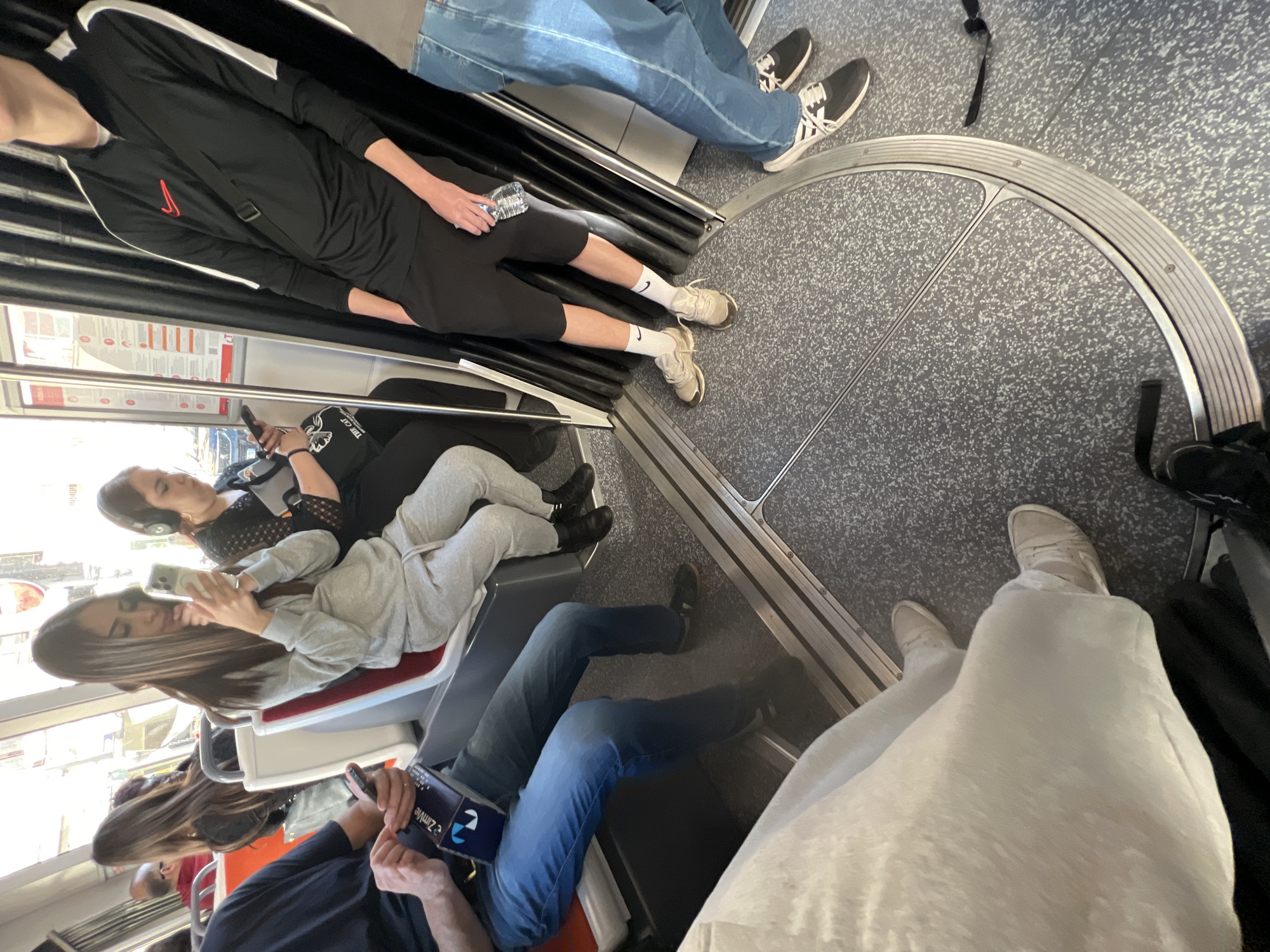 The image depicts the interior of a modern tram in Strasbourg, France. Several individuals are visible, seated or standing. At the top, two people wearing jeans with casual footwear are seen. A person in a black tracksuit with white socks and trainers holds a water bottle. Further down, two people are seated, looking at their phones. One is wearing headphones. Below, another person in jeans is looking at a Zimvie branded object. The floor of the tram is patterned gray. Metal railings and handholds are present. The tram windows reveal glimpses of the outside environment. At the bottom right, a person in white pants and beige trainers stands with a bag.